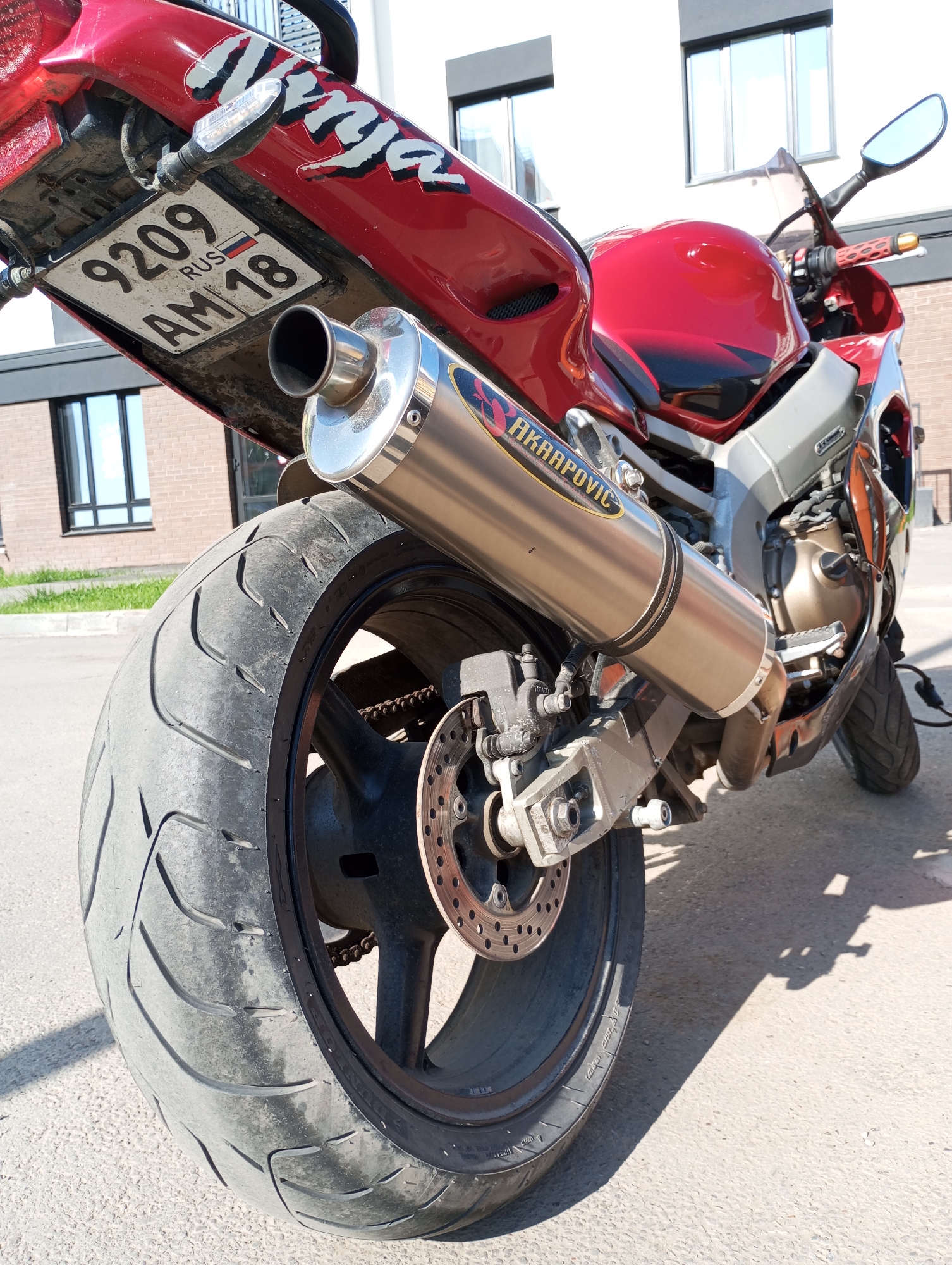 The image shows the rear of a red "Ninja" motorcycle parked outdoors. The motorcycle features an Akrapovic exhaust pipe and a Russian license plate that reads "9209 AM 18". The tire appears worn. The location may be in the city of Pervomaysky, Russia. In the background, there is a building with windows, suggesting an urban or suburban environment. The scene is captured in daylight.