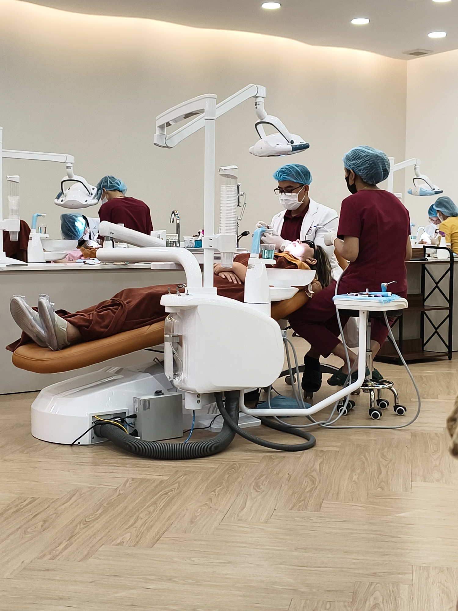 The image depicts a dental clinic with a patient in a dental chair. A dentist, wearing a white coat, mask, and surgical cap, is examining the patient's mouth. A dental assistant in maroon scrubs sits on a rolling stool nearby. Dental equipment surrounds the patient and dental staff. In the background, there are additional dental chairs and staff attending to other patients. The clinic has white walls, bright overhead lighting, and a light-colored wood-patterned floor. Location: Unknown, Unknown.