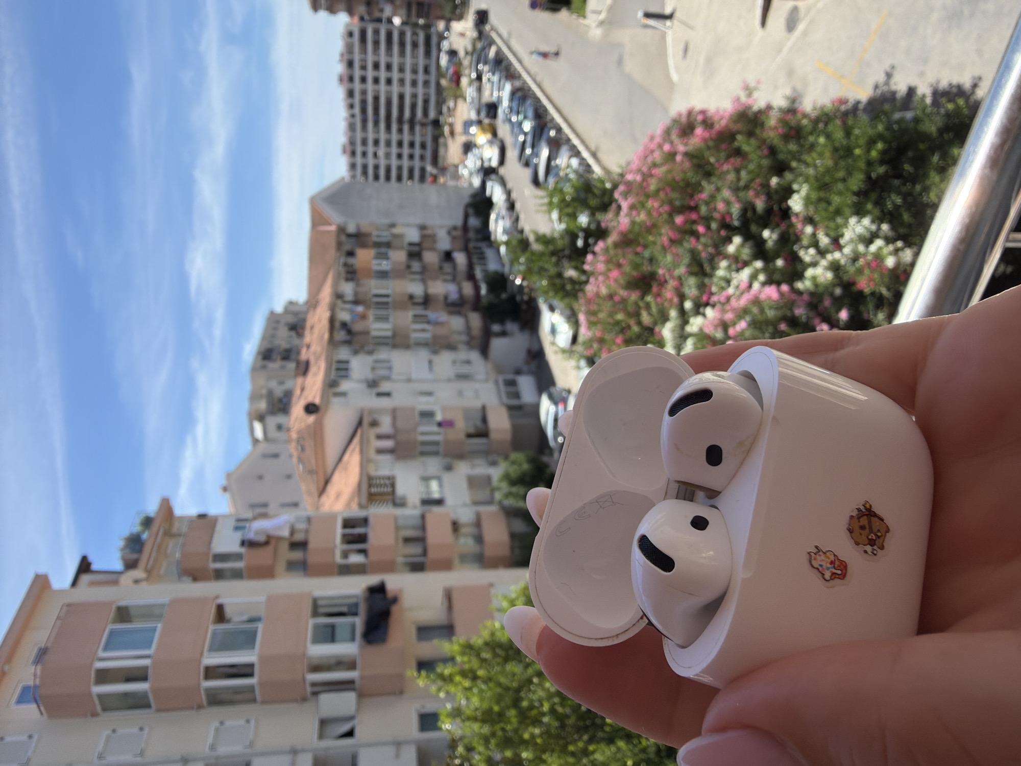 A close-up shot shows a person's hand holding a white wireless earbud charging case. Inside the case, two white earbuds with black accents are visible. The exterior of the case is adorned with two small stickers: one depicting a cupcake and the other a cartoon bear holding a bow and arrow. The background, captured from what appears to be a balcony in Budva, Montenegro, reveals a street lined with parked cars, apartment buildings, and lush green trees with pink and white flowers. The sky above is a clear blue with a few wispy clouds.
