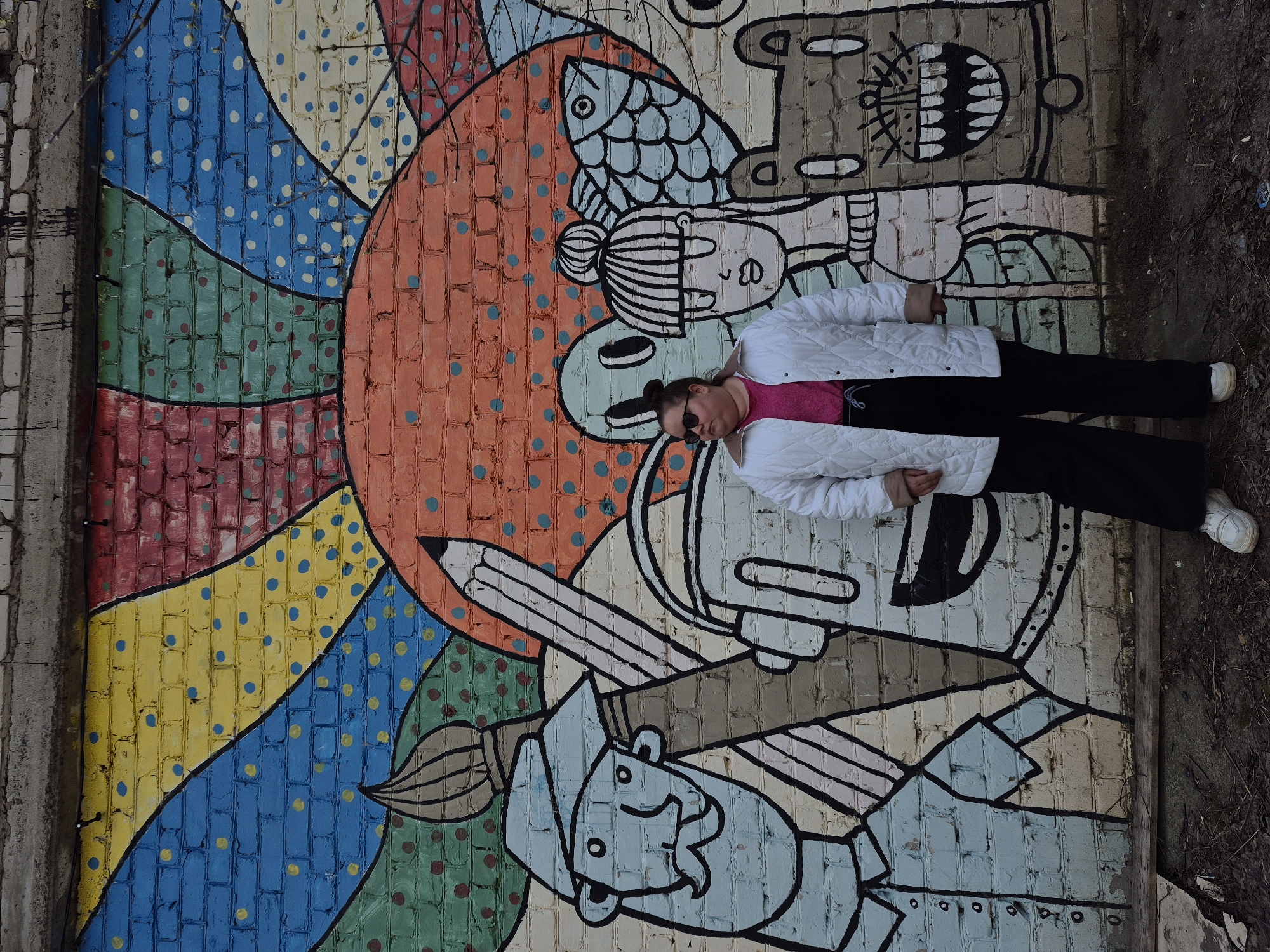 A young person is captured lying on the ground next to a brightly painted brick wall mural in Chaikovsky, Russia. The individual, dressed in a white quilted jacket, a vibrant pink top, black trousers, and white sneakers, wears sunglasses and appears to be posing playfully alongside the artwork.
The mural itself is a rich tapestry of color and whimsical design. On the left, a large, stylized sun-like form is depicted with radiating sections of blue, yellow, green, and red, each adorned with contrasting polka dots. To the right, various cartoon-like figures emerge from the vibrant background. These include a character with a fish on its head, another seemingly holding a large pencil, and a collection of stylized faces and abstract shapes, all outlined in bold black lines. The mural’s style is reminiscent of comic art or graphic novels.
The setting appears to be an outdoor urban space, likely a side street or alley, given the uneven, muddy ground visible to the right of the person. Bare tree branches, subtly visible in some areas, suggest a cooler season, possibly late autumn or early spring. The soft, diffused lighting indicates an overcast day or the gentler light of dawn or dusk. The overall image portrays a creative and relaxed interaction between the person and their colorful surroundings. No text is visible on the mural or elsewhere in the image.
