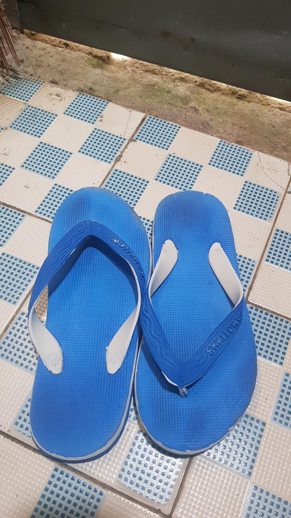 A pair of blue flip-flops with white straps are positioned side-by-side on a tiled floor in Bakalan, Indonesia. The floor features a pattern of white squares interspersed with blue checkered squares. Behind the flip-flops, the ground is visible, covered with dirt and small rocks, with a wooden plank or structure running horizontally across the top of the image. One of the flip-flops has the embossed text "SWALLOW" on the strap.