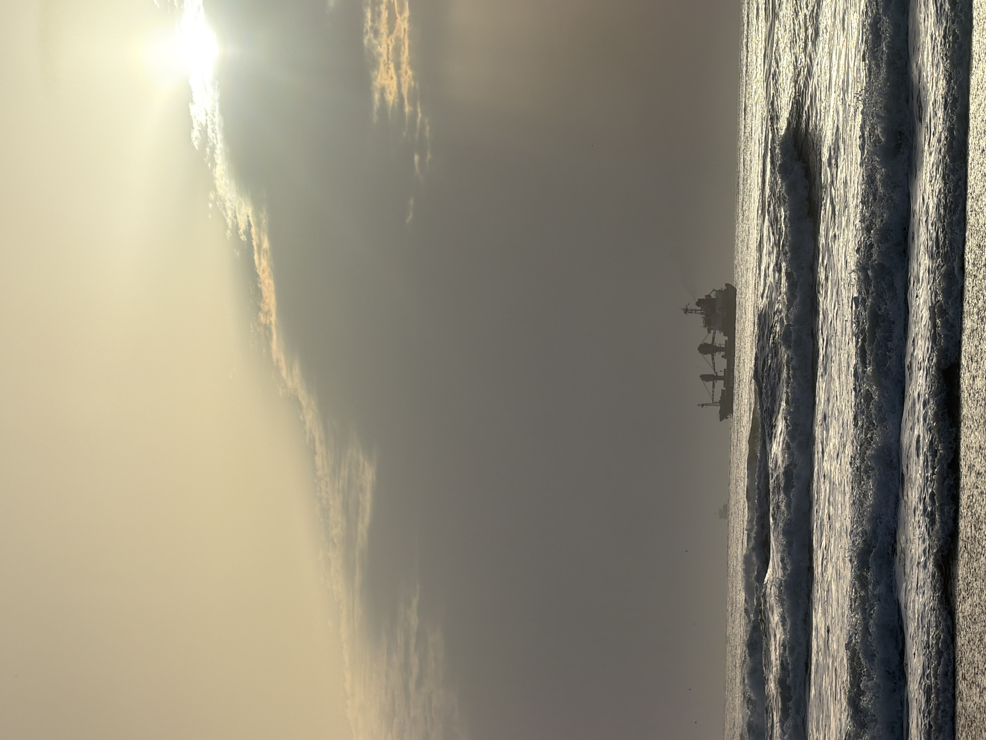 The image shows a beach scene, likely near Figueira da Foz, Portugal. Strong sunlight breaks through the clouds on the left side of the frame. The sea occupies the right side of the frame, with visible waves. A large ship is silhouetted in the distance on the horizon.