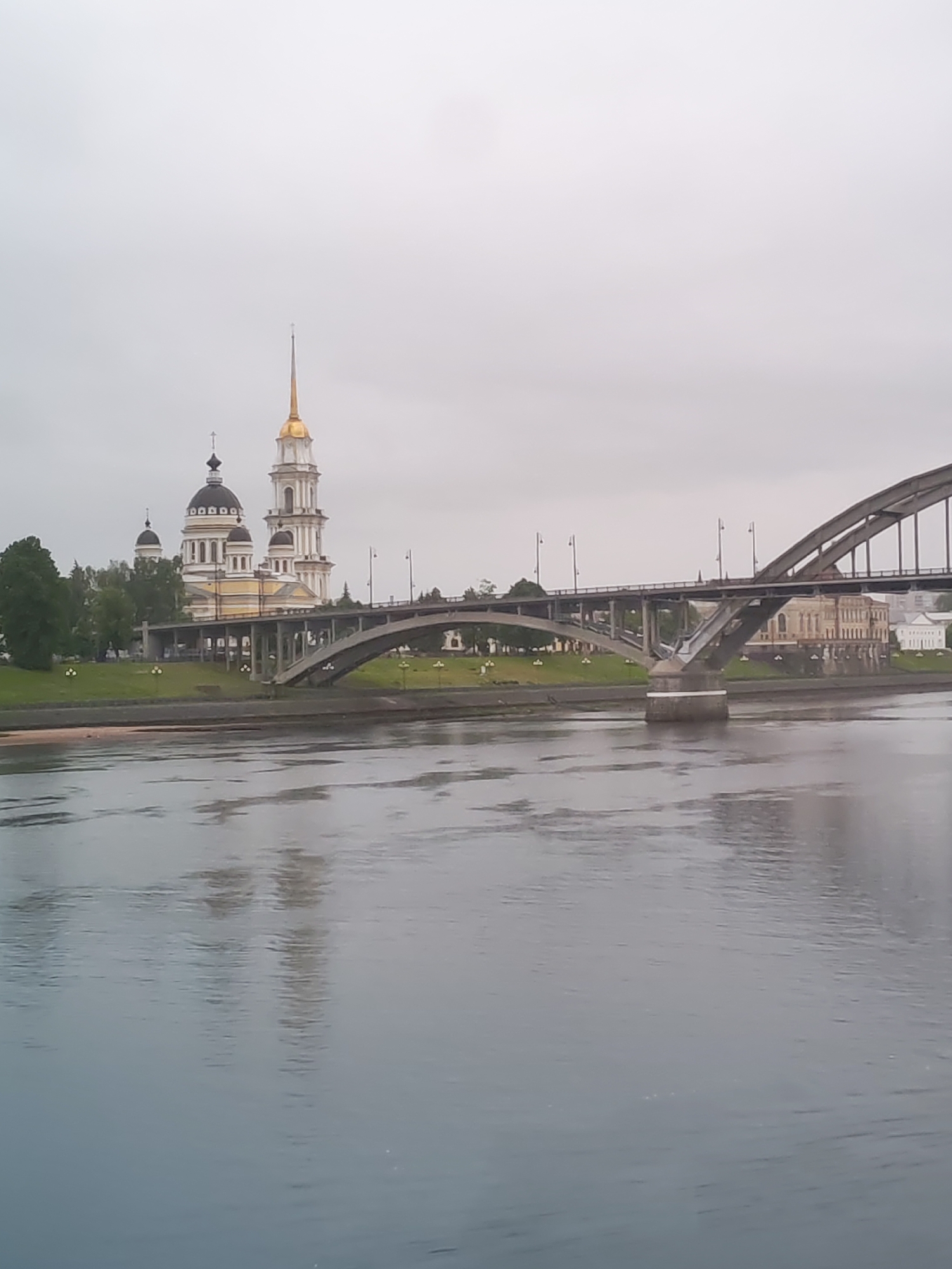 The image depicts an urban landscape in Bol'shie Mkhi, Russia, showcasing a wide river reflecting the overcast sky. A bridge arches across the river, supported by concrete pillars. On the riverbank, a large cathedral-like building with a golden spire and black domes stands prominently next to a grassy area lined with streetlights. The photograph captures a serene, somewhat muted atmosphere due to the cloudy weather.