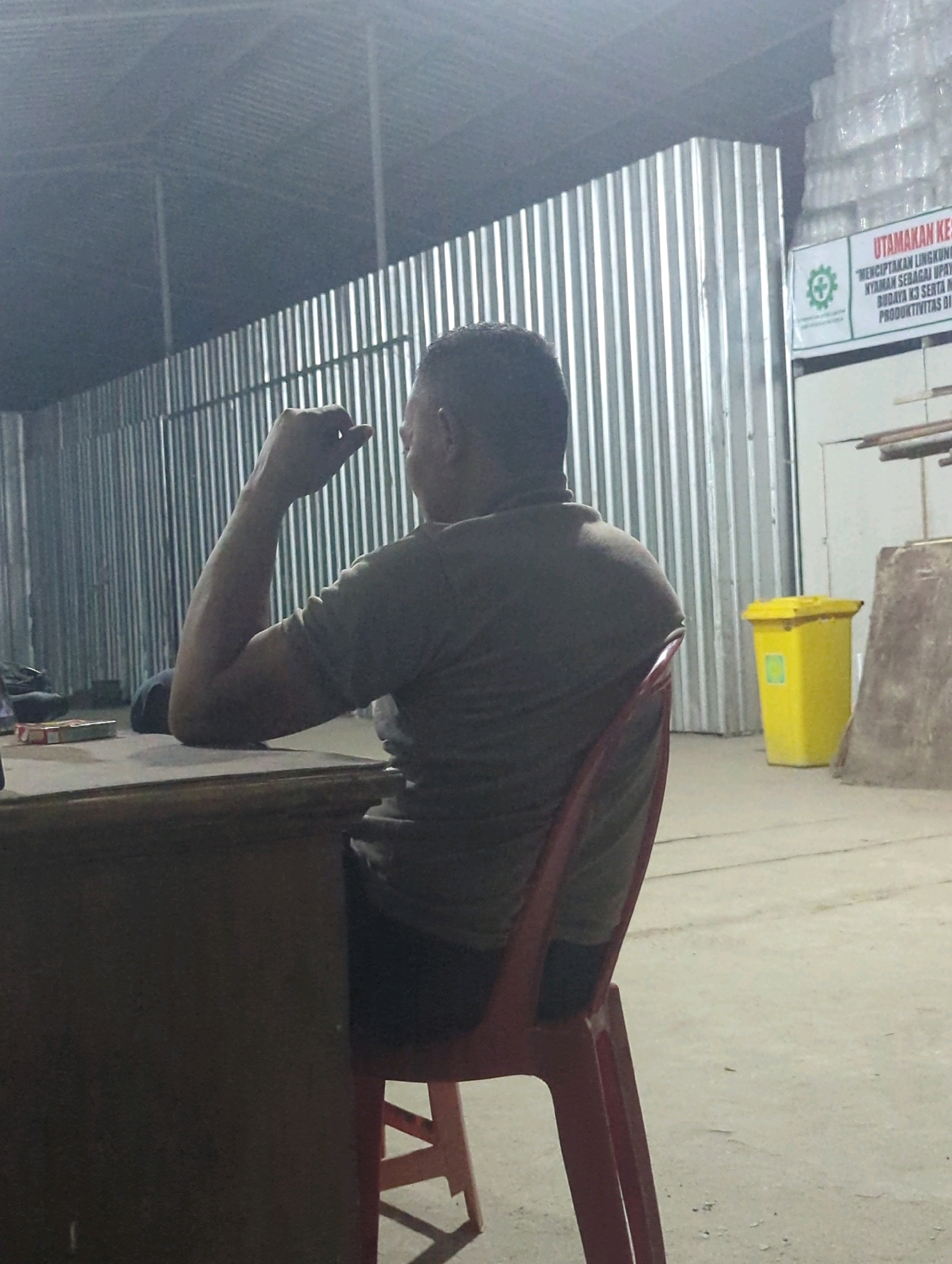 The image depicts the interior of a warehouse-like structure, possibly in Batu Belaman, Indonesia. A man is seated from the back, leaning on his arm and looking towards a corrugated metal wall. He is wearing a dark grey t-shirt and dark pants. In front of him is a wooden table. To his right, a red plastic chair is visible. In the background, a yellow trash bin with a green logo is positioned against the metal wall. A sign with text in Indonesian is mounted on the wall to the right of the trash bin. The floor appears to be concrete. The lighting is dim, suggesting an evening or indoor setting with artificial illumination.