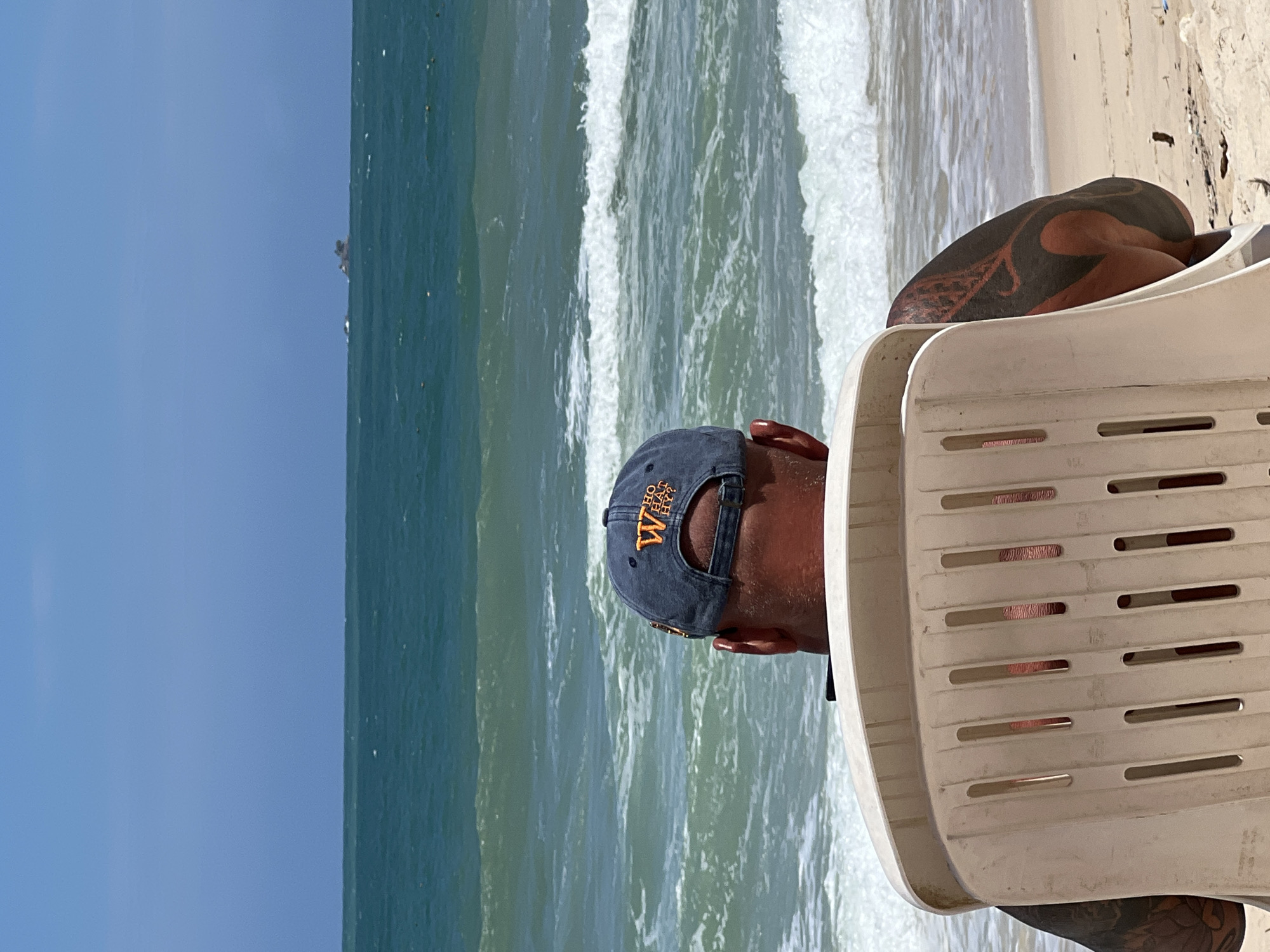 In Choeng Thale, Thailand, a person sits in a white plastic chair facing the ocean. They are wearing a dark blue baseball cap with an orange logo that reads "WHO WHAT WHY?". The person has extensive black tattoos on their arms and back, visible through the slats of the chair and on their exposed shoulder. The ocean is turquoise with white-capped waves rolling towards a sandy beach. In the distance, a small island with vegetation is visible on the horizon under a clear blue sky. The scene is a candid outdoor shot capturing a moment of relaxation by the sea.