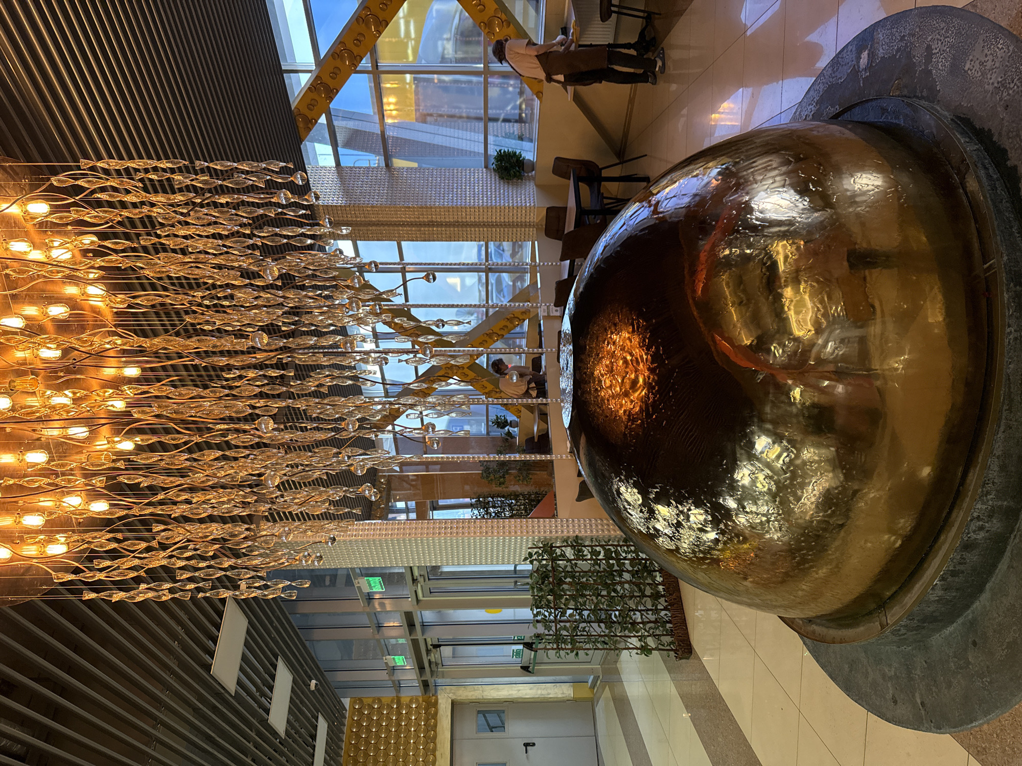 The image shows the interior of a modern building with a large, golden, spherical sculpture placed on a gray, tiered base. A chandelier composed of multiple light bulbs and glass elements hangs from the ceiling on the left side of the frame. Large windows are visible in the background, revealing a cityscape with a distinctive yellow structure. The floor is tiled in light beige. In the background, there are some plants along the windows. A person wearing a white shirt and dark pants is working in the background. A door with an "exit" sign is visible.
There is no information to suggest the location is in the city of Petropavlovsk-Kamchatsky, Russia.