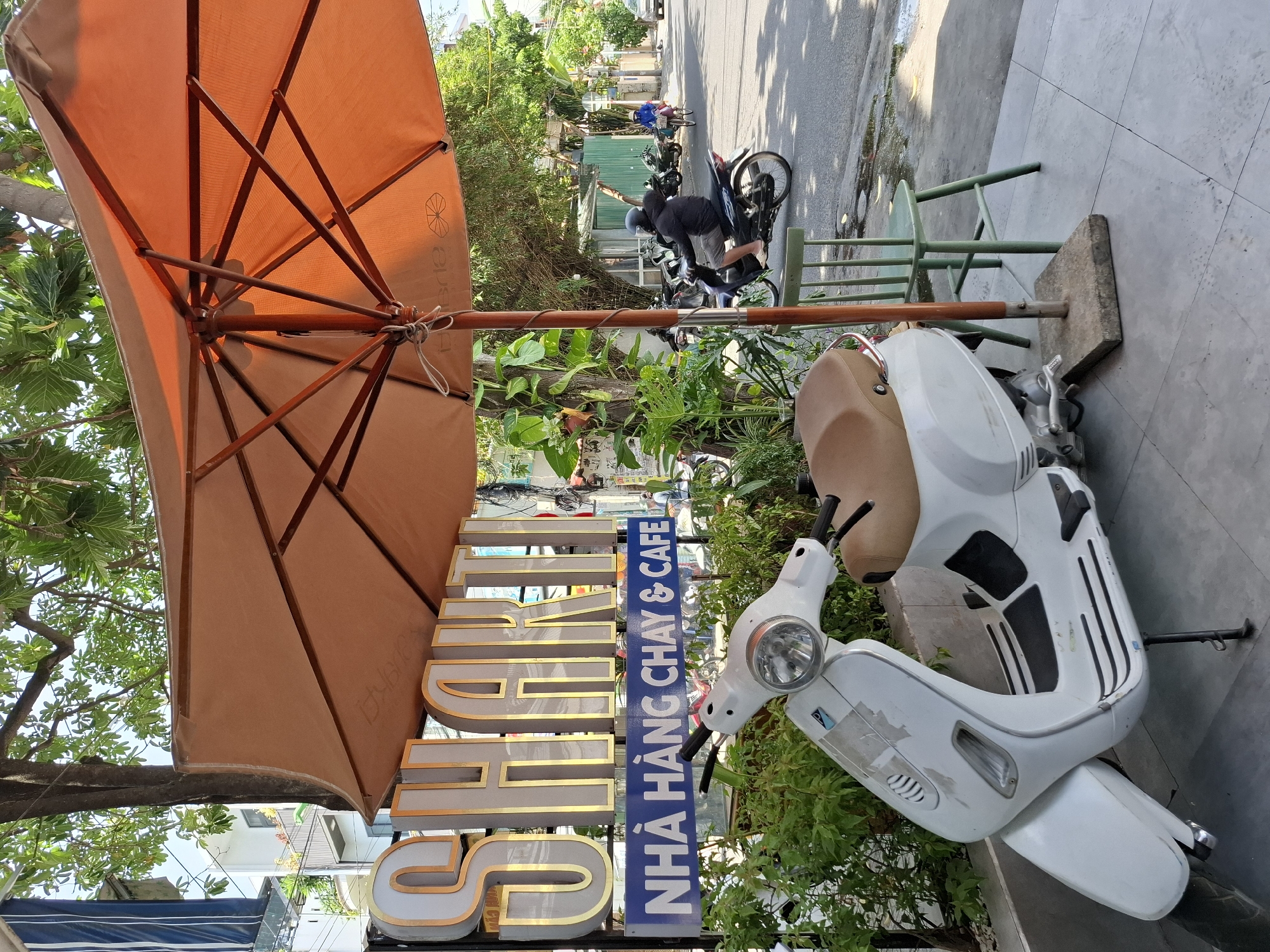 This is a medium shot, eye-level, outdoor photograph taken in Nha Trang Ward, Vietnam. The foreground features a white Vespa scooter parked on a grey tiled patio. The scooter is facing left, with its kickstand down. Behind the scooter and to the right, a green wooden chair is positioned. A large orange umbrella with a wooden pole, anchored by a concrete block, extends into the frame from the top. In the background, a street scene unfolds with a few motorcycles and a person on a bicycle. The building behind the scooter has a prominent sign that reads "SHAKTI" in gold lettering, with a blue sign below it displaying "NHÀ HÀNG CHAY & CAFE" in white lettering. Lush green foliage surrounds the signage and the patio area. The overall impression is a street-side cafe setting.