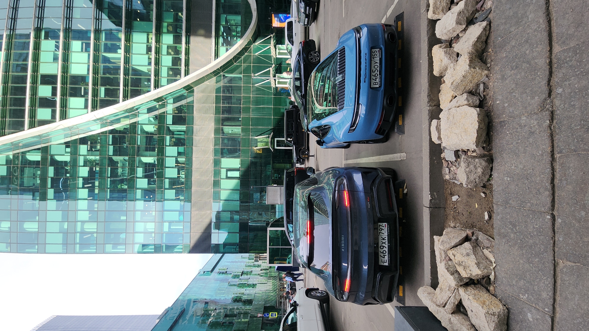 A ground-level, outdoor shot depicts parked cars in front of modern buildings. The architecture is notable for its extensive use of glass. The vehicles include what appears to be a blue sports car and a dark sedan, identified by the "Xiaomi" badge on its rear. The buildings are in Moscow, Russia. The parked cars and pedestrians are visible in the reflecting building. A stack of large rocks is visible on the right. The sky is visible in the upper left.