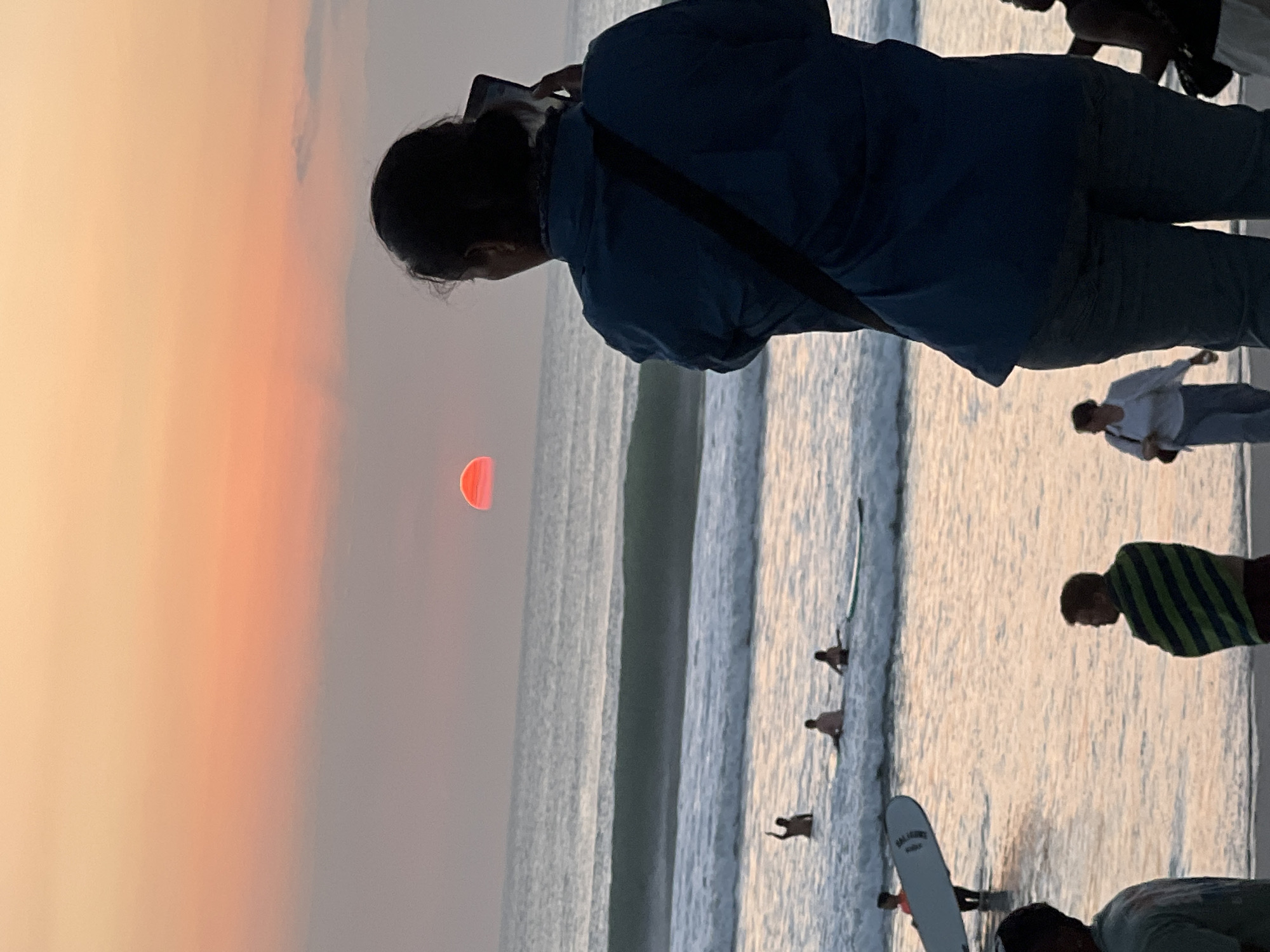 The image depicts a sunset scene at Kuta Beach, Bali, Indonesia. In the foreground, a person with dark hair pulled back is standing and taking a photo of the sunset with their phone. They are wearing a blue shirt and grey pants. To the left and in the mid-ground, several people are in the ocean, some with surfboards. One person is standing with their arm raised, and others are sitting on surfboards in the water. Further to the left, a person is holding a surfboard. In the background, the sun is low on the horizon, casting a reddish-orange glow across the sky and reflecting on the water. Waves are breaking on the shore. Other beachgoers are visible in the foreground and mid-ground, some walking and others standing. The overall atmosphere is that of a relaxed beach setting during sunset.