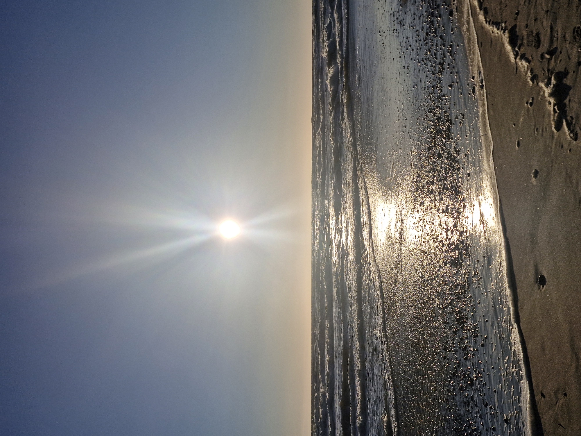 The image shows a sunset over the Baltic Sea at the Curonian Spit in Klaipėda, Lithuania. The sun is low in the sky, casting a warm glow on the water and the beach. The waves are gentle, and the sky is clear and blue. There are no people or animals visible in the image. The foreground is dominated by the wet sand of the beach, with small pebbles scattered across its surface. The wet sand reflects the sunlight, creating a glittering effect. The middle ground is occupied by the sea, with lines of small waves rolling towards the shore. The horizon line is clearly defined where the sea meets the sky. The background is a clear, unblemished sky, transitioning from a lighter hue near the horizon to a deeper blue towards the top of the frame. The sun is a bright, luminous orb positioned in the upper center of the image, emitting prominent rays of light that fan out across the sky and reflect on the water.