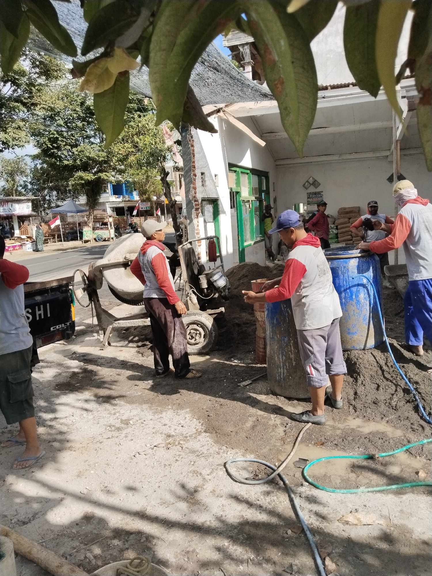In Prigi, Indonesia, several construction workers are actively involved in mixing cement. A concrete mixer is positioned next to a truck. Piles of cement are visible, along with large blue barrels. One worker is using a shovel to move cement, while another is preparing to add material to a barrel. The scene is outdoors, with a building in the background. The ground is dusty and uneven, with some puddles of water. The overall impression is of a busy construction site under a clear sky.