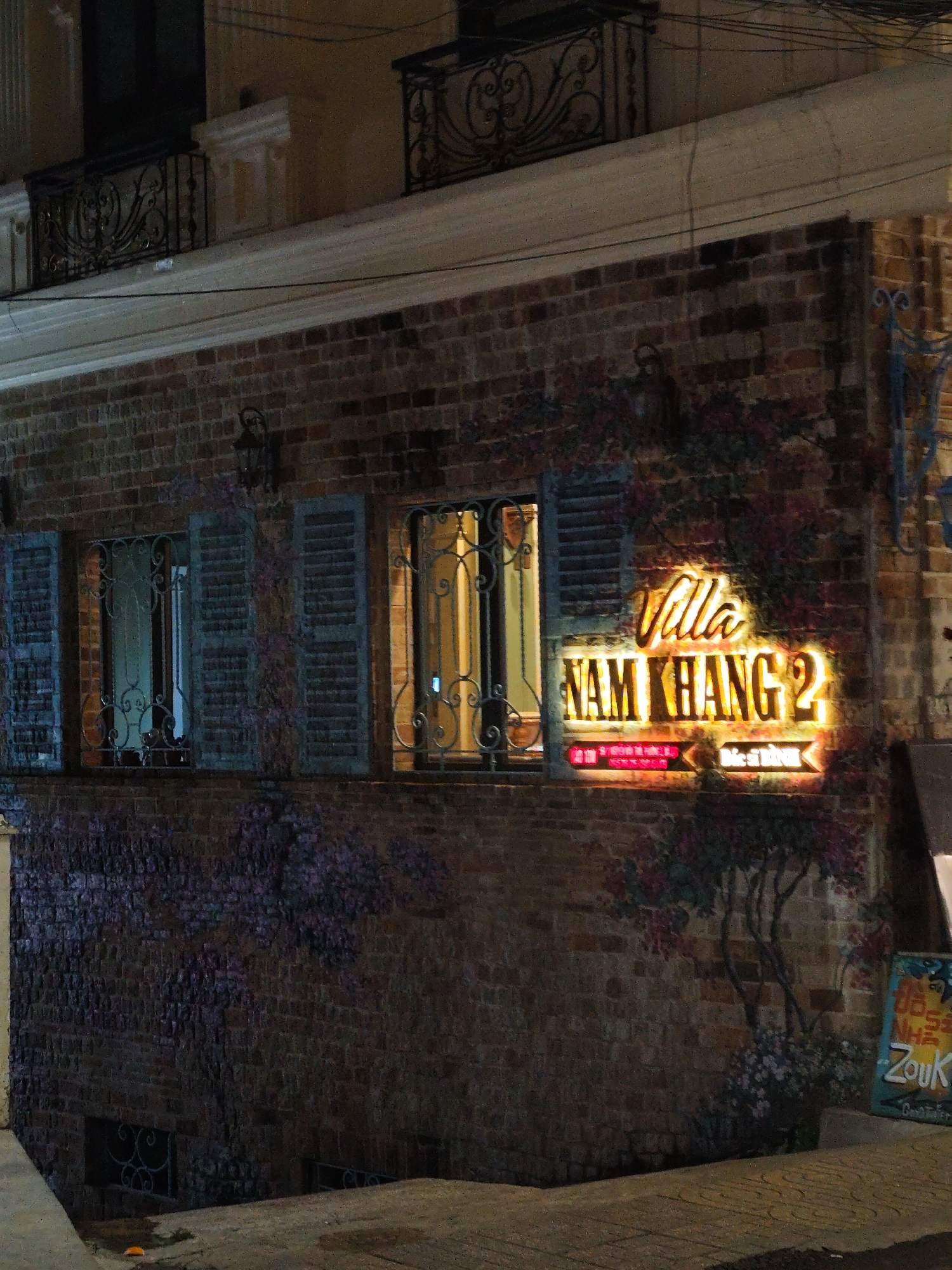 The image depicts the exterior of "Villa Nam Khang 2" in Da Lat, Vietnam, illuminated by a neon sign. The building is brick with shuttered windows and decorative wrought iron. A mural of purple flowers is painted on the brickwork. A sign with "Zouk" is visible next to the entrance. The scene is captured at night.