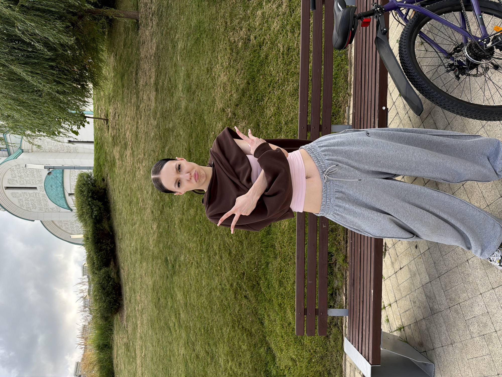A young woman with dark hair pulled back in a ponytail stands in front of a park bench. She is wearing a brown cropped hoodie over a pink tank top and oversized gray sweatpants. Her arms are crossed in front of her chest, and she is making a pouty face. She is standing on a paved area with a bicycle parked to her right. Behind her, there is a grassy hill with a building and trees. The building is white with a green dome and appears to be a church or similar structure. The location is a park in the city of Krasnogorsk, Russia.