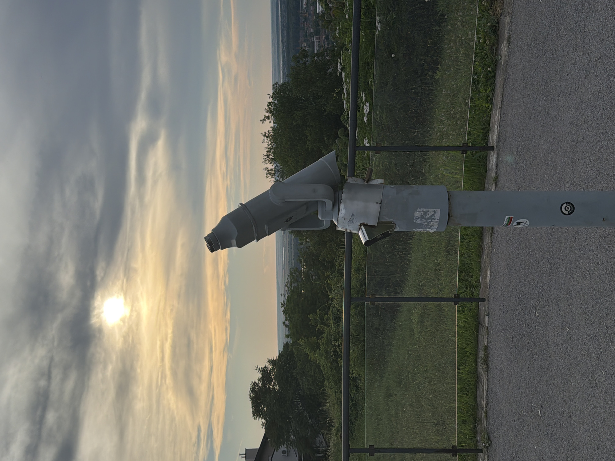 The image depicts a viewpoint overlooking the city of Ruse, Bulgaria, likely from a higher elevation. In the foreground, a grey, coin-operated public telescope is positioned on a grey metal pole. The telescope is pointed towards the horizon, which is slightly hazy and shows the city and a body of water. The sky above is partly cloudy with the sun visible, partially obscured by clouds. The immediate surroundings include a paved pathway in the foreground, a grassy slope with a low metal railing and glass barrier, and dense green trees and vegetation behind the railing. A building with a light-colored facade and air conditioning units is visible on the left, further up the slope. The overall scene suggests an observation point offering a panoramic view of the city and its surroundings.