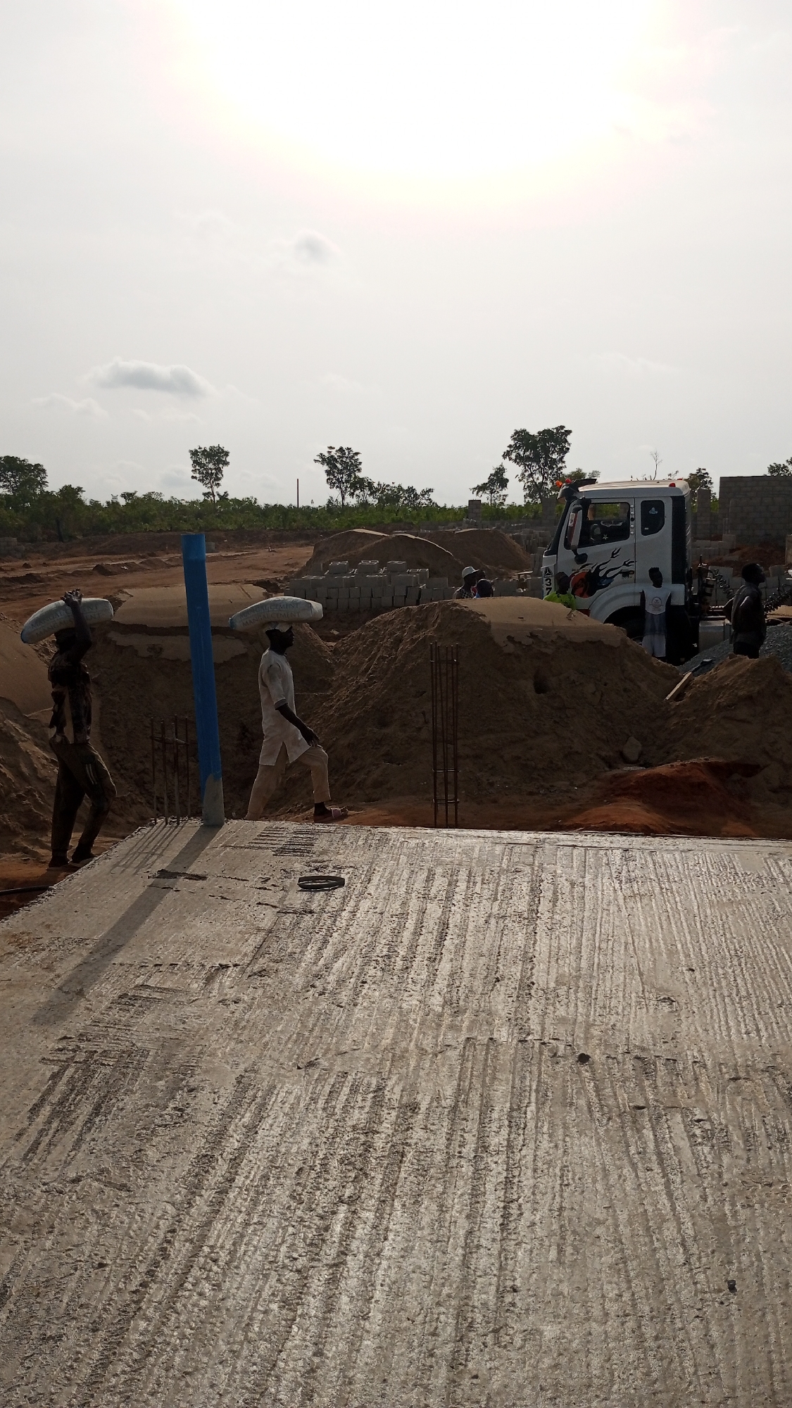 The image shows a construction site in Zango-ndaji, Nigeria. Two men are carrying bags on their heads. A truck is parked near a pile of sand and concrete blocks. Reinforcement bars are visible, protruding from a partially constructed concrete slab. Other construction workers are visible in the background. The environment includes trees and a cloudy sky.
