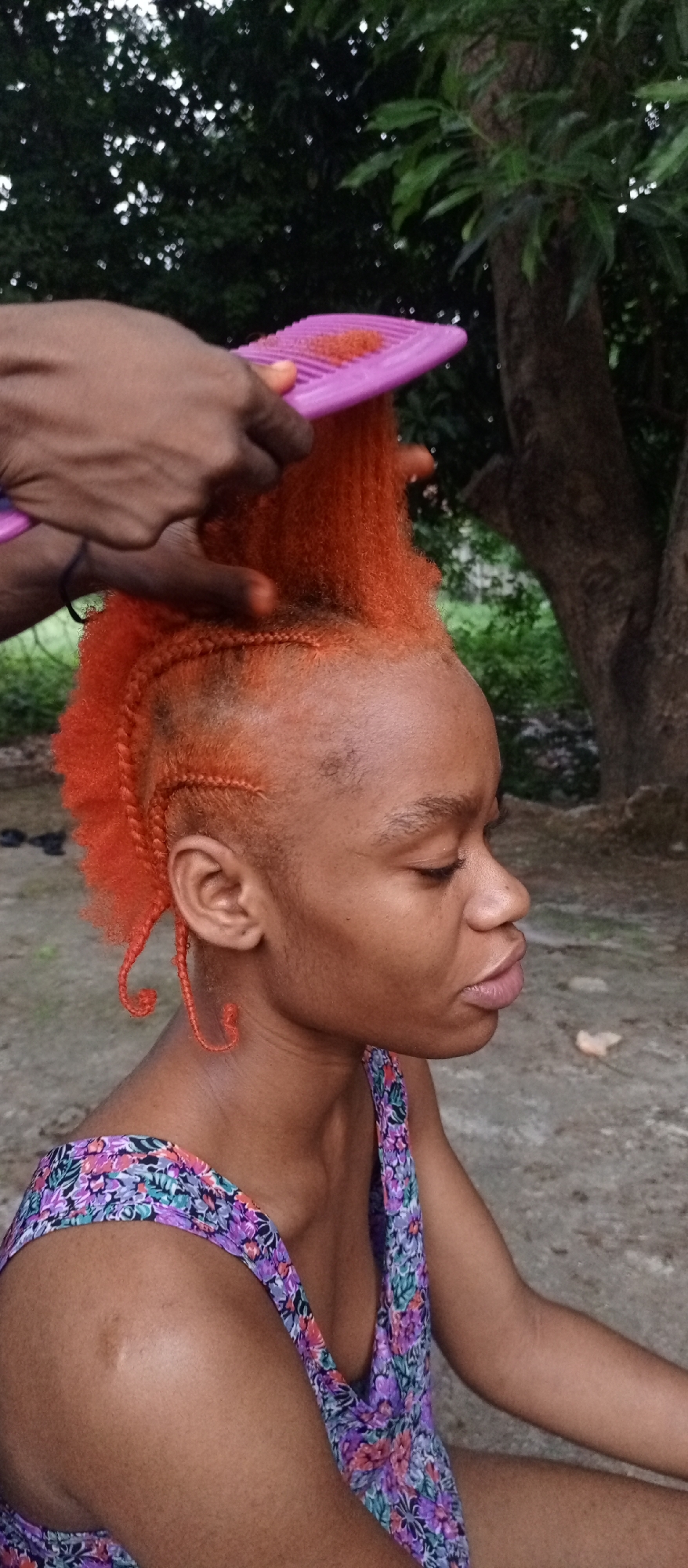 A woman is having her orange hair styled with a purple comb outdoors in Keffi, Nigeria. The woman has cornrows along the side of her head with curled ends. She is wearing a floral print top and is looking down with her eyes closed. Another person is holding the comb and styling her hair. A tree is visible in the background. The setting appears to be a casual outdoor environment.