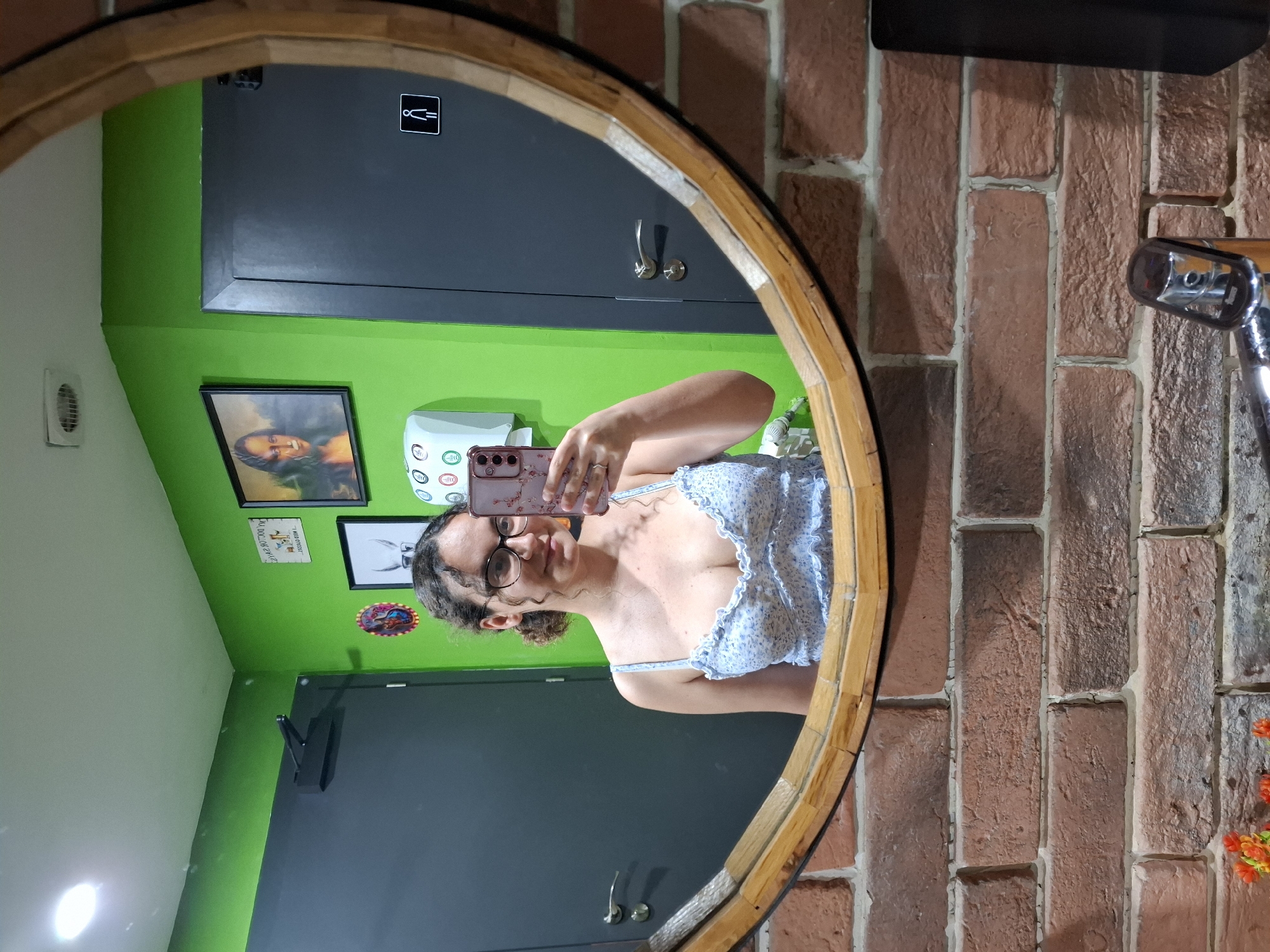 A woman with curly brown hair and glasses takes a mirror selfie in a restroom. She is wearing a light blue floral camisole. Behind her, the wall is painted bright green, and there is a framed print of the Mona Lisa and a restroom sign. The mirror is round with a wooden frame, and the lower portion of the image shows a brick wall and a sink faucet.