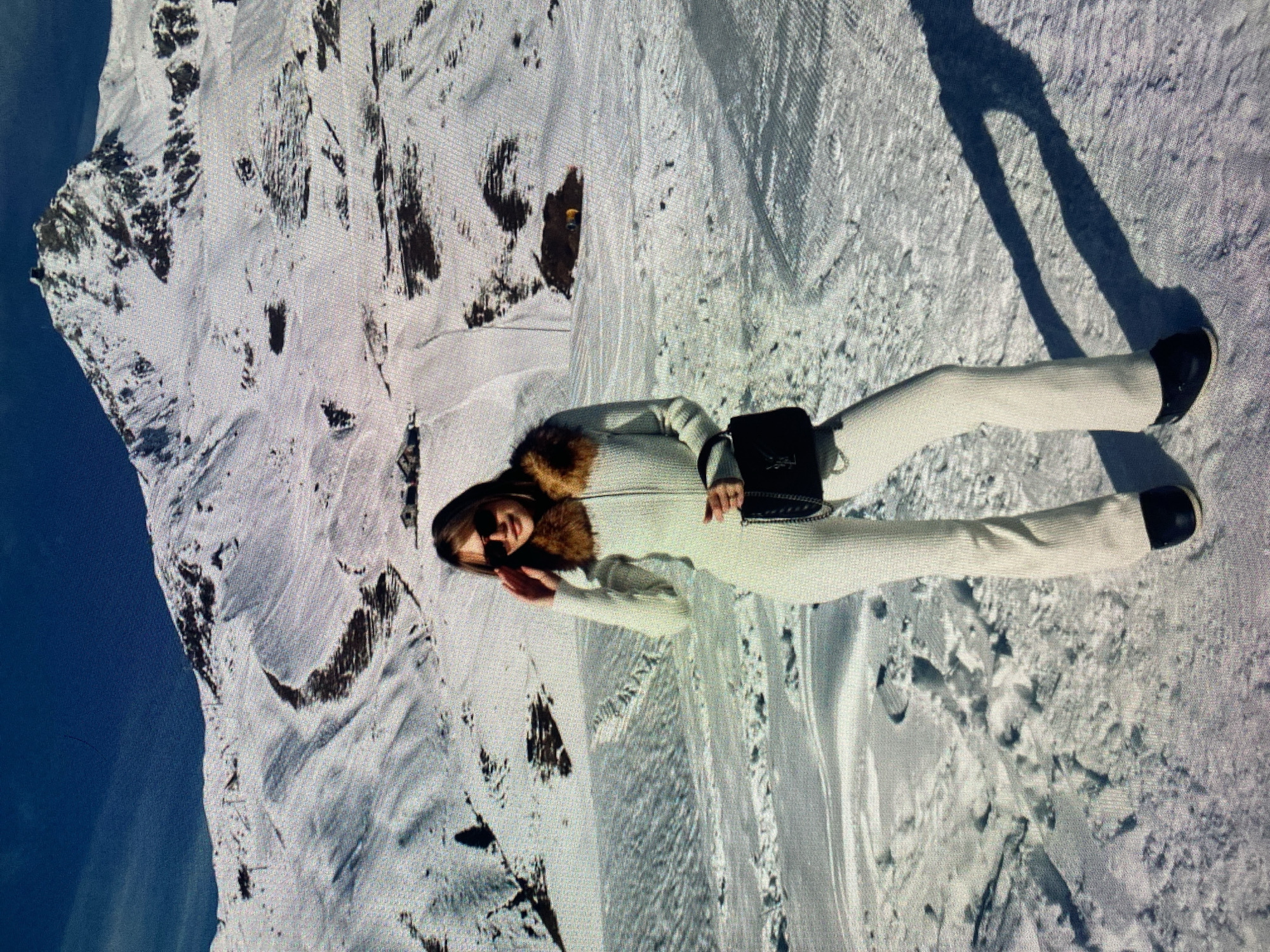 A person wearing a white snowsuit with a fur collar, sunglasses, and black boots is lying on a snowy mountainside. They are holding a hand to their face. The mountain is covered in snow, and there are dark patches of rock or vegetation visible. A shadow of a person taking the photo falls on the right side of the image. The image is rotated 90 degrees clockwise. The location is not in Łódź, Poland.