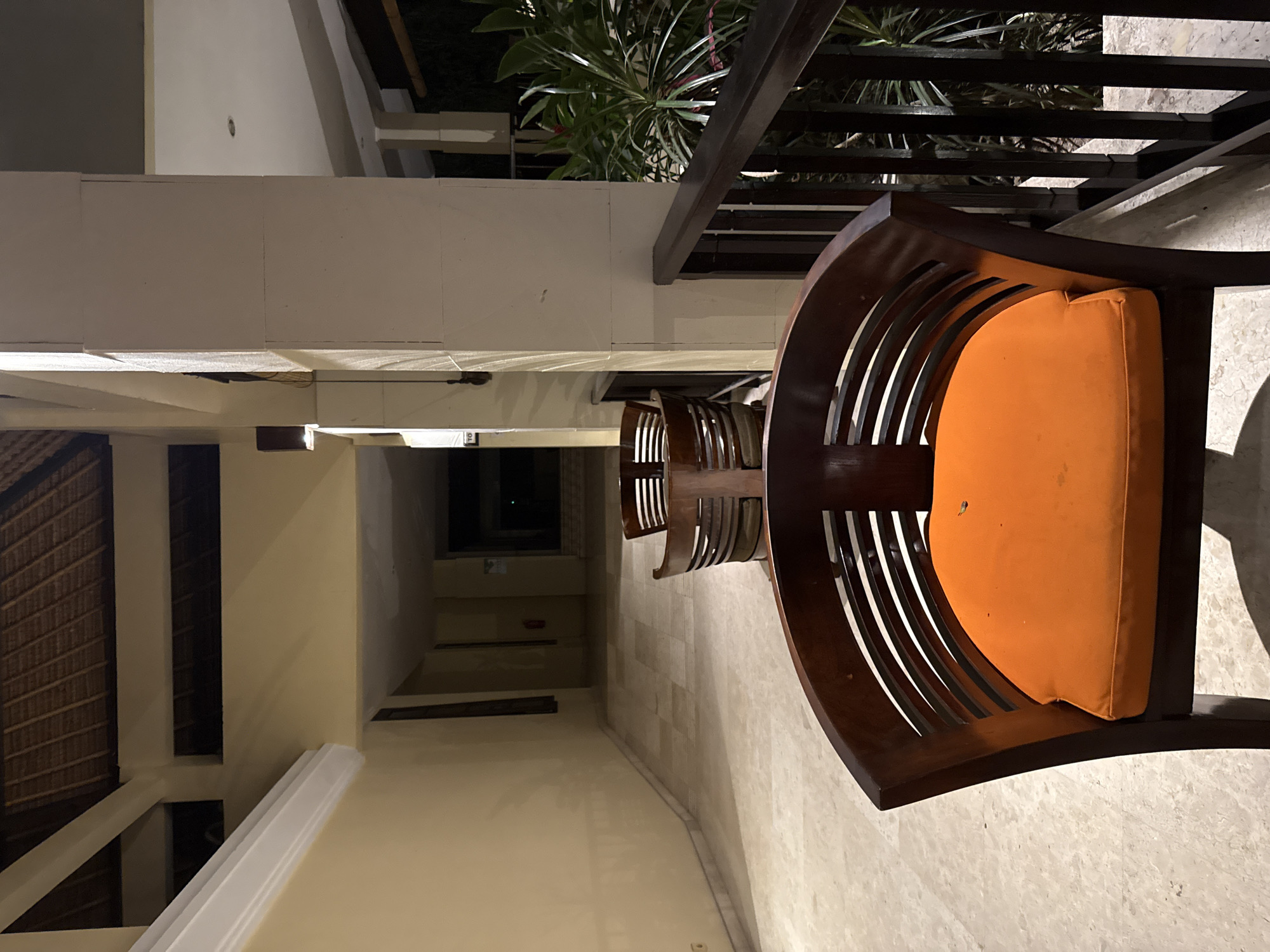 The image depicts an outdoor seating area at night, likely a resort or hotel, in Denpasar, Indonesia. In the foreground, a dark wooden chair with an orange cushion is visible, with another similar chair partially obscured behind it. These chairs are arranged along a railing overlooking a lush green plant. The background shows a dimly lit hallway with a tiled floor and cream-colored walls. A thatched roof is visible on the ceiling of the walkway. The scene is devoid of any people or discernible activity.