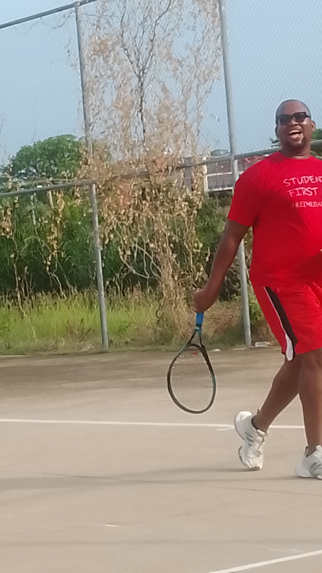This outdoor image captures a joyful man engaged in what appears to be a tennis match or practice session in Lokoja, Nigeria. The man, with dark skin, is wearing dark sunglasses, a bright red short-sleeved t-shirt, and matching red shorts with a black and white stripe on the side. He is in motion, with his right foot slightly lifted and his body leaning forward, holding a tennis racket with a blue grip in his right hand. His face is expressive, beaming with a wide, open-mouthed smile, conveying palpable happiness and excitement.
On his red t-shirt, white text is clearly visible, reading "STUDENT FIRST" followed by "REDHEAD" below it. He is wearing white athletic shoes. The setting is an outdoor court with a light-colored paved surface and a white line marking the playing area in the foreground. Behind him, a chain-link fence separates the court from a natural background featuring trees and shrubs. Some foliage is lush and green, while a large tree directly behind the man appears dry or dormant with light brown branches and leaves. The sky is clear and light blue, indicating bright, sunny weather, likely mid-day or afternoon. The scene evokes a sense of active enjoyment and leisure.