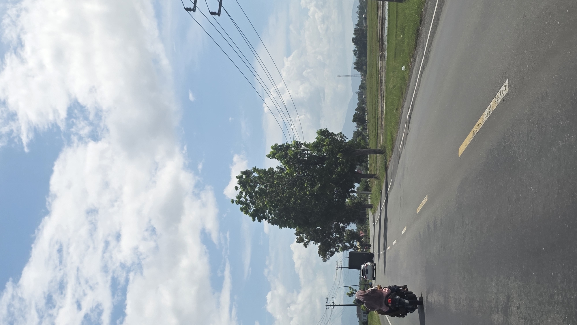 The image shows a road in Kampungjumpa, Indonesia, with a motorcycle driving on it. The road has a yellow centerline and is lined with trees and grass. In the background are power lines and a cloudy sky. A few cars are also on the road. The image appears to be taken from a moving vehicle.