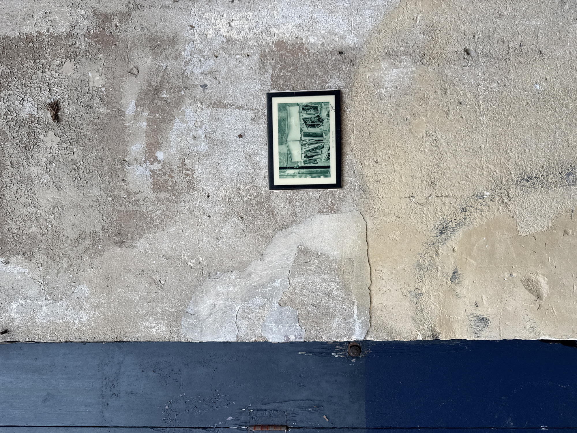 The image shows a section of a weathered, textured wall with peeling paint, predominantly in shades of grey and beige, adjacent to a dark blue wooden surface. A small, framed photograph, rendered in a teal-toned monochrome, is affixed to the wall. The photograph depicts a close-up view of a display of books or similar items on shelves. The overall setting appears to be an outdoor urban environment, suggesting a side of a building or an alleyway. The photograph's inscription at the bottom reads "3 janvier 2018 @graffitiphot". This detail, combined with the urban texture, suggests the photograph was taken in a city, possibly in the context of street art or ephemeral installations, consistent with a location like Bordeaux, France. No people are visible in the image.