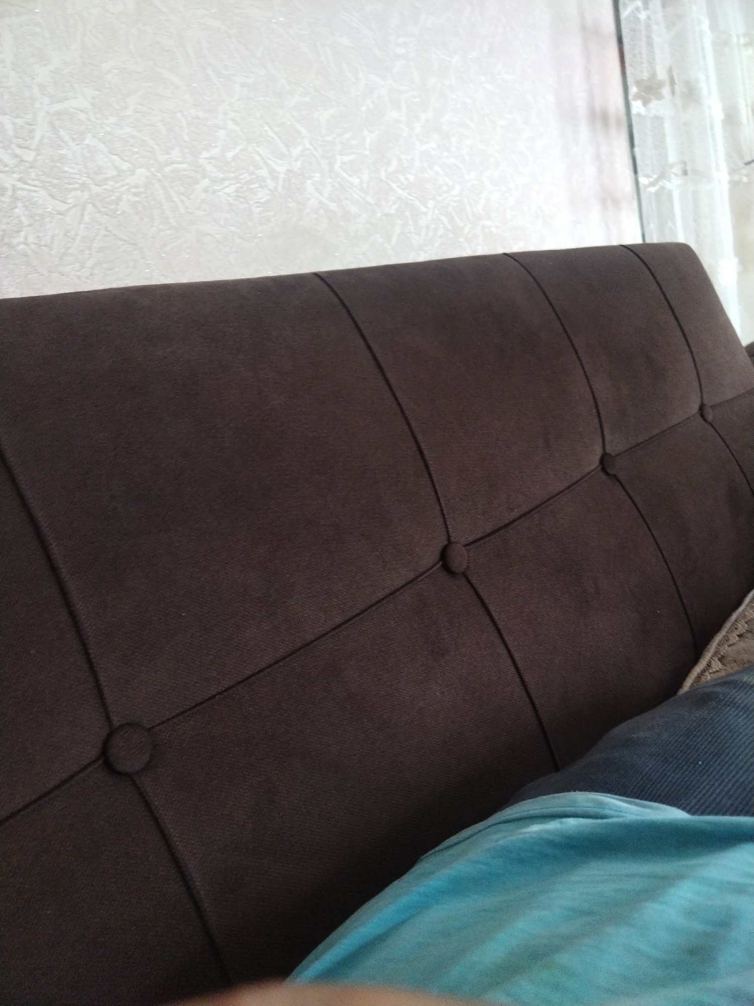 The image shows a close-up view of a dark brown tufted sofa in what appears to be an indoor setting. The sofa has a diamond pattern created by stitching and buttons, with two buttons clearly visible in the frame. The background consists of a textured off-white wall and a sheer white curtain. In the foreground, a bright turquoise-colored garment, possibly a t-shirt or blanket, is partially visible. There are no people or animals in the frame. The scene is well-lit, suggesting daytime or good artificial lighting. The overall impression is one of domestic comfort. No specific location in Khando, Georgia can be determined from the image.