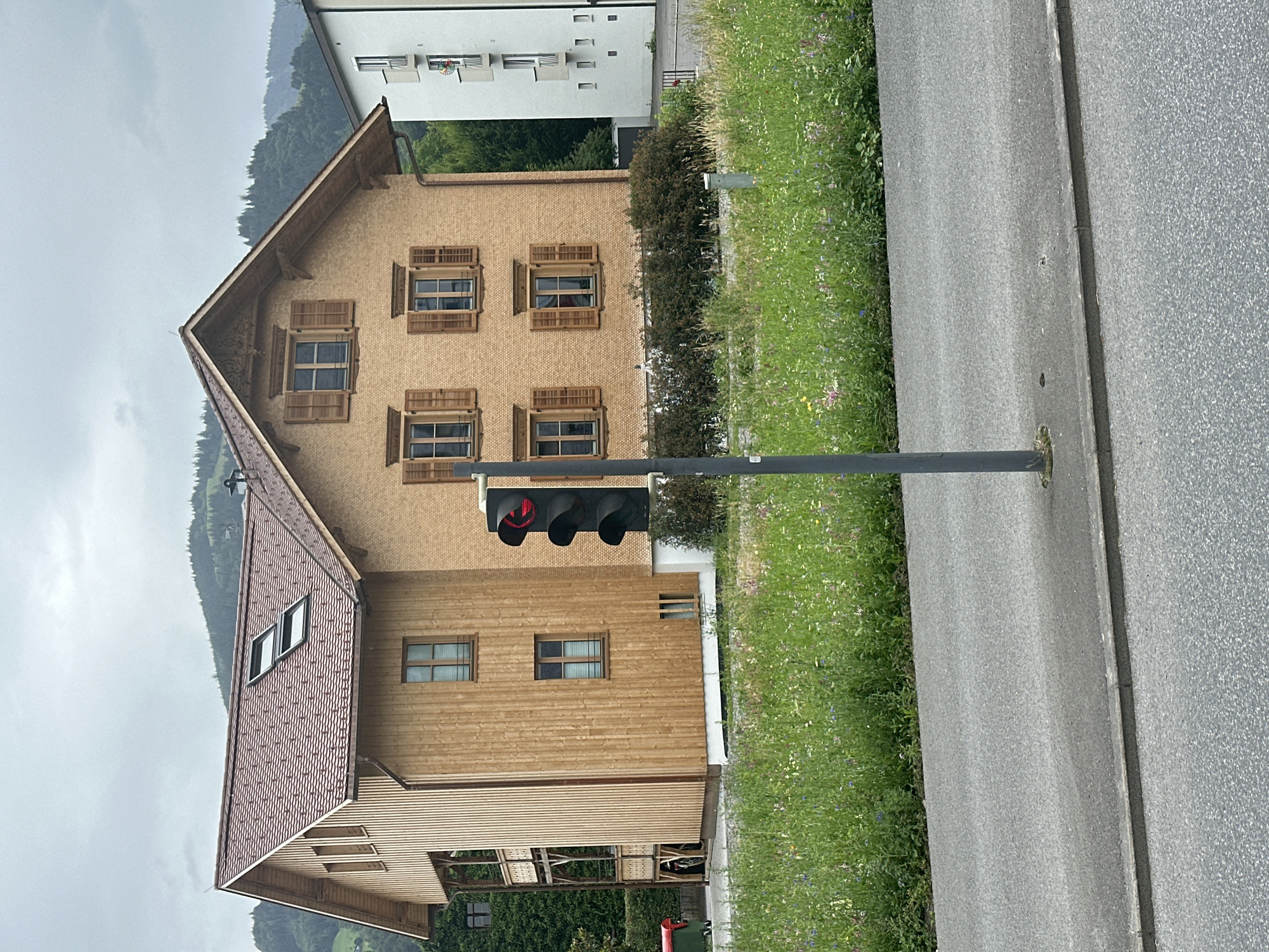 The image depicts a street scene in Dornbirn, Austria, featuring a multi-story wooden house with a red tiled roof. The house has multiple windows with wooden shutters. In the foreground, a traffic light is visible with a red light illuminated. Beyond the house, a modern white building stands. The area is characterized by a paved road, a grassy area with wildflowers, and a background of forested hills under an overcast sky. There are no people or animals visible in the scene.