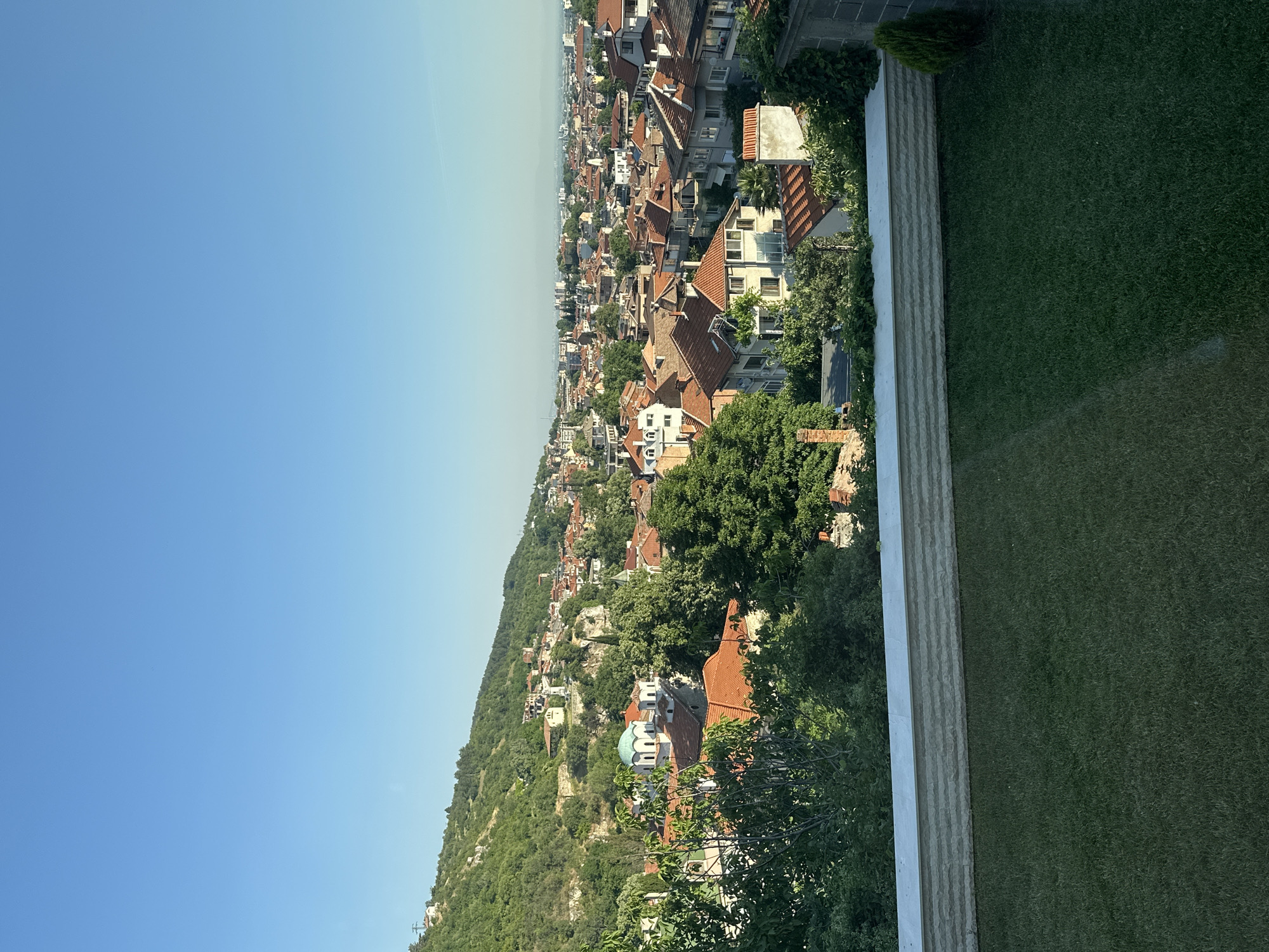 The image depicts an elevated, panoramic view of a city nestled against a verdant, hilly landscape. The foreground features a manicured green lawn and a low, textured stone wall. Beyond the wall, a dense canopy of trees partially obscures a collection of buildings with red tile roofs, characteristic of traditional architecture.
Further into the scene, the city unfolds with a multitude of houses, many exhibiting similar terracotta roofing. A prominent structure with a green dome, possibly a church or religious building, stands out amongst the rooftops. The city sprawls across the terrain, ascending the gentle slopes of the hills. The background reveals a more distant urban expanse with taller buildings, and the horizon is met by a clear, bright blue sky, indicating a sunny day. The overall impression is a serene, lived-in urban environment integrated with its natural surroundings. This view is highly characteristic of Asenovgrad, Bulgaria, known for its hilly terrain and historic architecture.