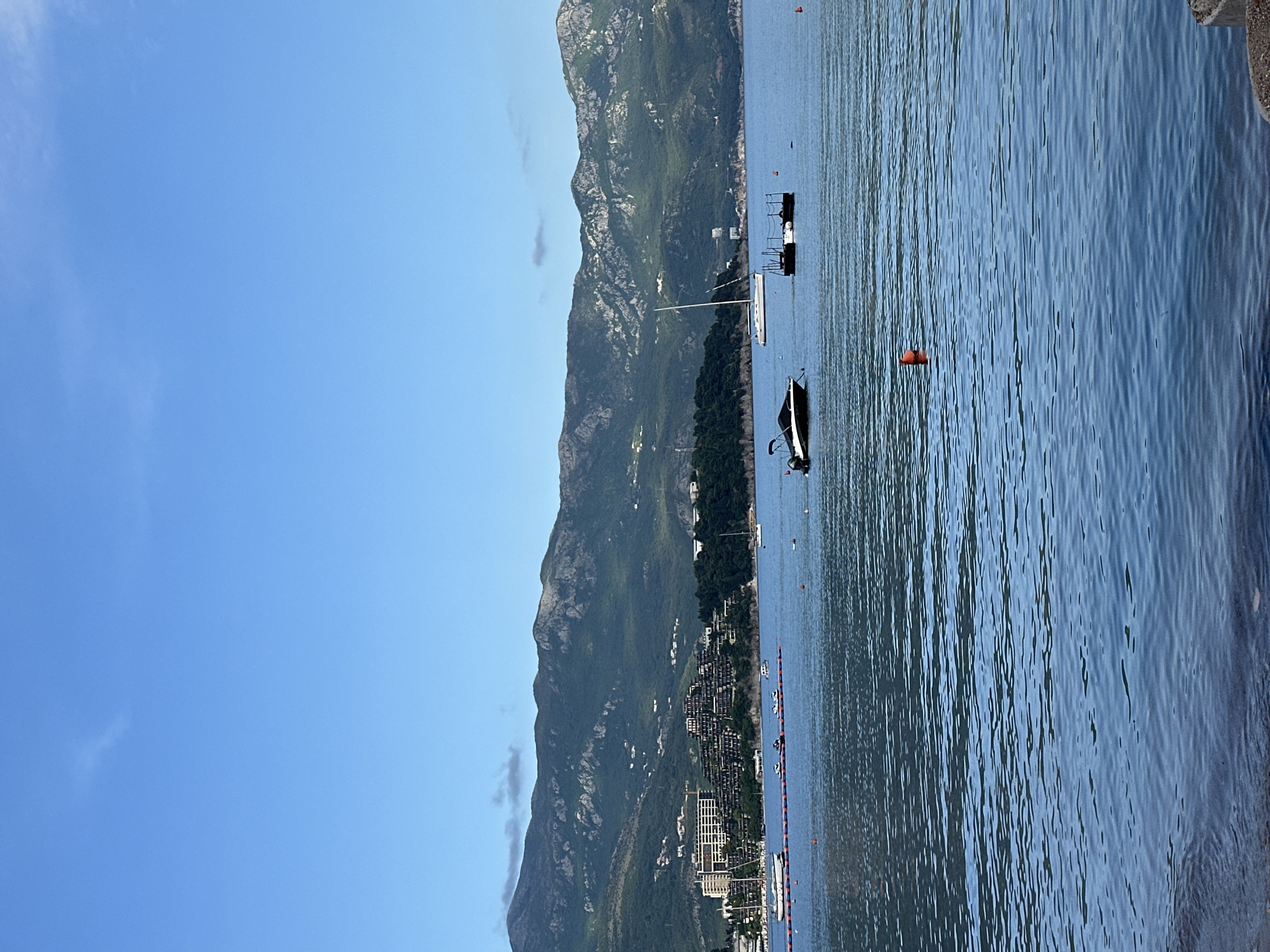 The image presents a view of Budva, Montenegro. The sea is visible with multiple boats anchored. A mountainous landscape rises behind the city. The sky is clear and blue with a few clouds. Buildings line the shoreline. Orange buoys are in the water.