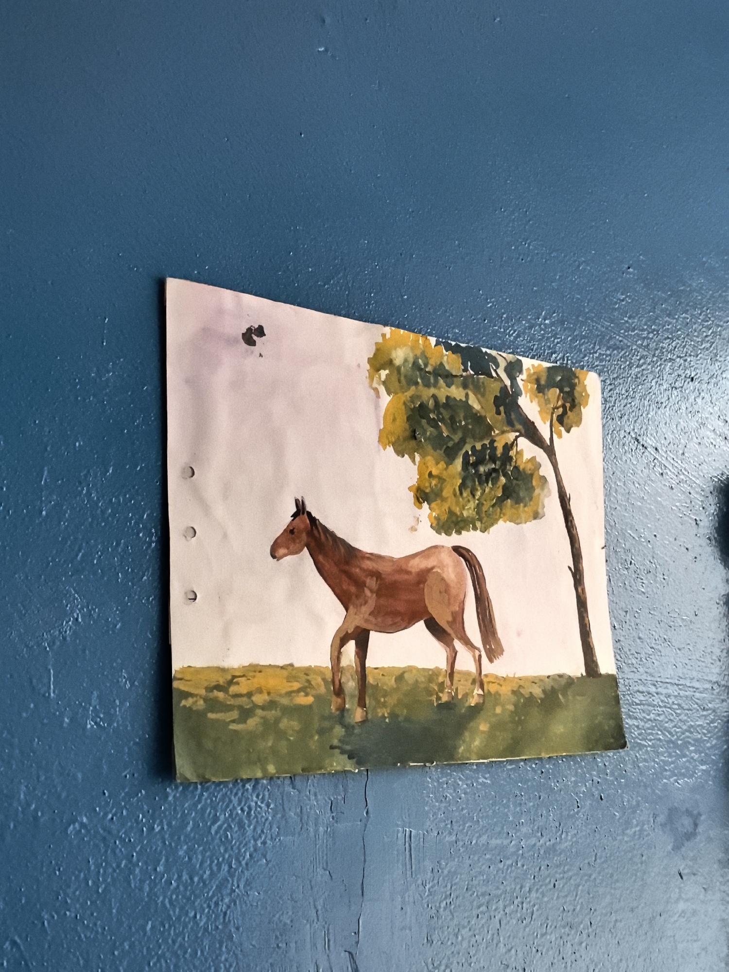 An unframed, watercolor painting of a brown horse standing in a grassy field with a tree is affixed to a textured, blue wall. The painting is positioned in the center of the frame, angled slightly downwards to the right. The horse is depicted in profile, facing left, with its head held high. The tree has green and yellow foliage and a brown trunk. The grass is painted in various shades of green and yellow. There are three circular holes punched along the left edge of the painting, suggesting it was once part of a binder. The wall's surface is rough and uneven, with visible paint textures and some minor imperfections. The lighting suggests the image was taken indoors, possibly in a room with natural light from an unseen source. The location is unknown, as it is not specified within the image or its context.