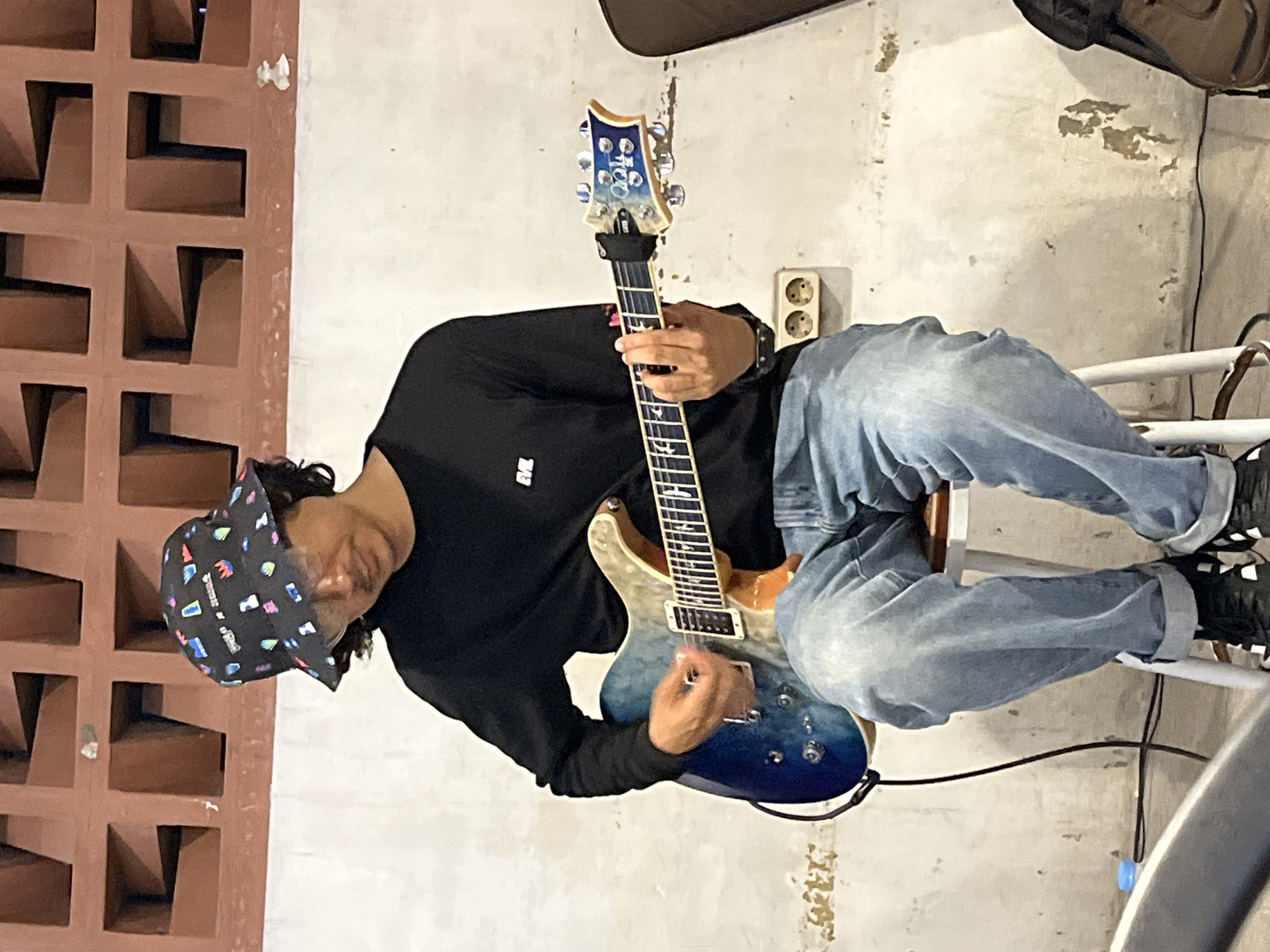 A man is playing an electric guitar while sitting in a room. He is wearing a black long-sleeved shirt, light blue jeans, a patterned bucket hat, and black shoes. He is seated on a white chair with metal legs. The guitar is blue and has a light-colored neck. A black cable is connected to the guitar and runs along the floor. Behind the man is a white wall with some peeling paint. A brown wall with square cutouts is to his left. An electrical outlet is visible on the white wall. A brown soft case rests against the wall behind him. The location is Lhokseumawe, Indonesia.
