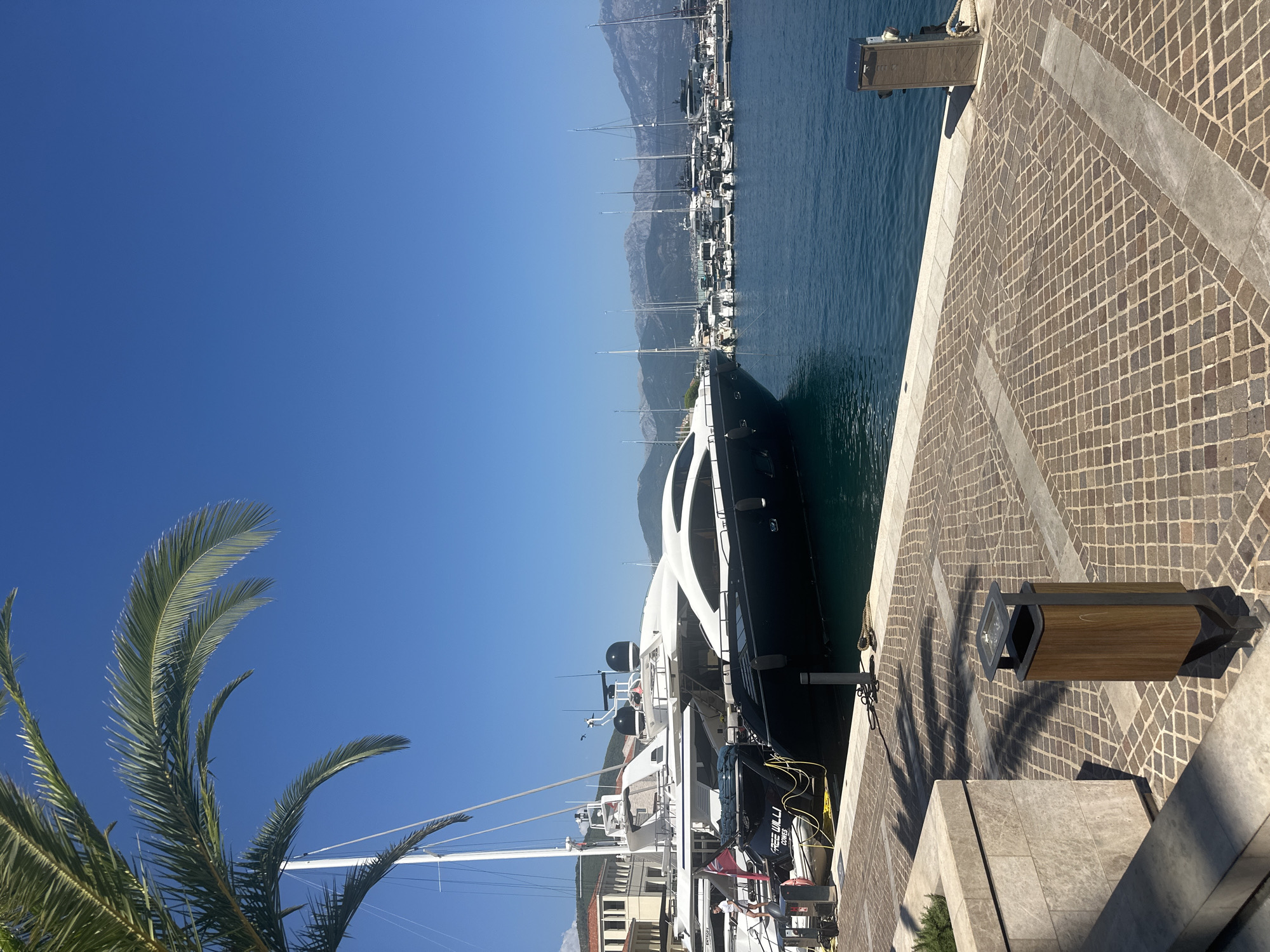 The image depicts a harbor in Herceg Novi, Montenegro, on a clear, sunny day. The foreground is dominated by a paved walkway with a stone border, featuring a modern wooden trash bin and the shadow of a palm tree cast upon it. To the left, a portion of a stone planter is visible.
The main subject is a large, sleek, black and white yacht docked in the foreground, its hull reflecting the bright sky. Adjacent to it, another white yacht is partially visible. In the background, a marina filled with numerous sailboats and other vessels stretches towards a mountainous landscape under a vivid blue sky. Buildings with terracotta roofs can be seen on the left side of the frame, suggesting a waterfront town. The water in the harbor is a deep blue, calm, and reflects the clear sky and the yachts. A single person, dressed in white, is standing on the stern of the black and white yacht, appearing to be interacting with equipment or preparing to disembark. Another smaller boat is tied to the side of the yacht. The overall scene conveys a sense of luxury and tranquility characteristic of a Mediterranean port.