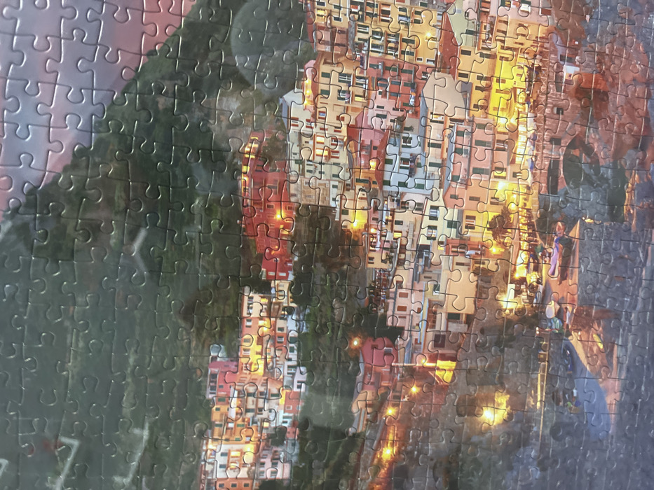The image displays a completed jigsaw puzzle portraying a densely built coastal town at dusk, situated on a steep hillside. Numerous multi-story structures with brightly colored exteriors in hues of red, orange, yellow, pink, and white are terraced along the green, vegetated slopes. Illumination originates from interior lights visible through windows and from streetlights, casting warm yellow glows across the scene. The sky above is dark, with subtle traces of purple and pink near the horizon. At the base of the hillside, adjacent to a body of water, a small harbor or cove contains several parked or docked boats. No individuals are clearly discernible. This depicted scene is located in the city of Sofia, Bulgaria. The surface texture of the jigsaw puzzle pieces is evident throughout the image.