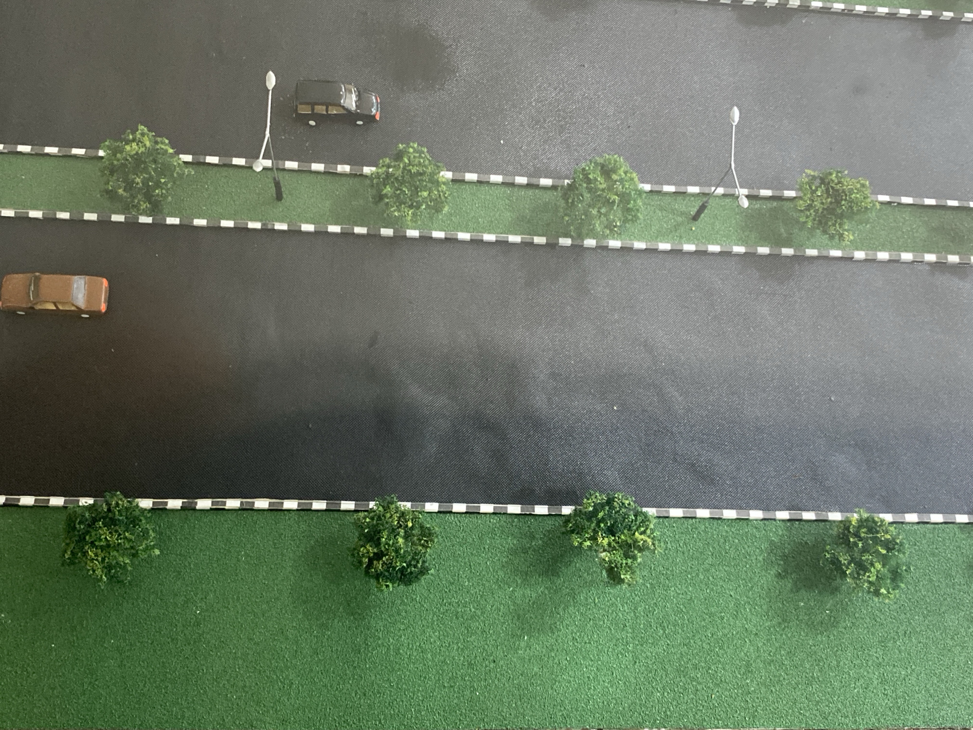 This is a top-down, medium shot of a miniature diorama depicting a city street. The diorama is comprised of a black asphalt road running horizontally across the frame, bordered by green grassy areas with small trees and decorative white and black striped edging. Two lanes of traffic are indicated by the positioning of toy cars. In the upper lane, a black SUV is parked, and further down, a white lamppost stands near a small green tree. In the lower lane, a brown car is in motion, and a row of small trees and lampposts are visible further down the street. The lower portion of the diorama shows a wider expanse of green grass with more small trees. The scene is static, with no discernible action or interaction. The environment appears to be a stylized representation of an urban or suburban street, likely a model for a railway or architectural display.