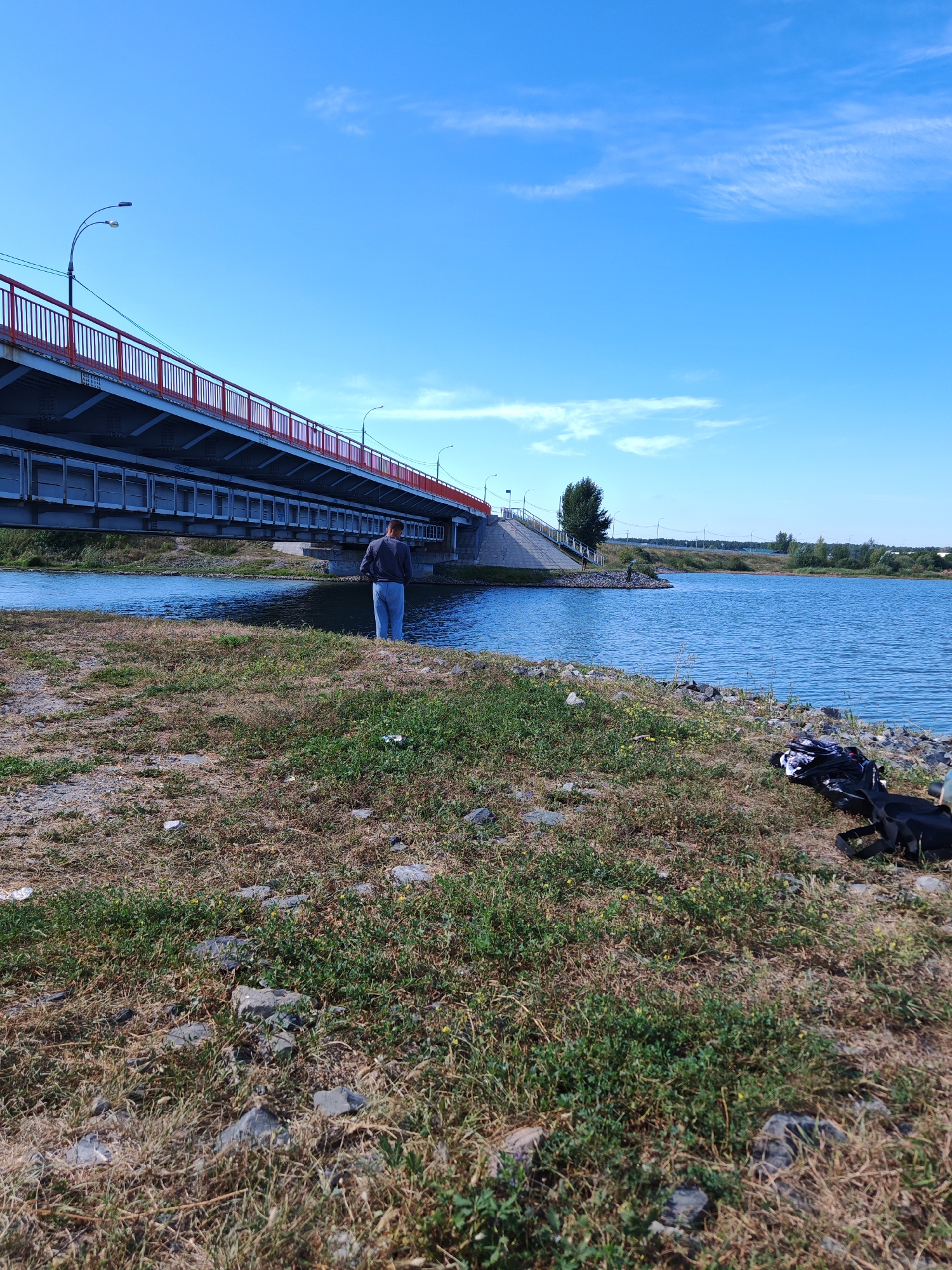 A full-shot medium angle, exterior, daytime photograph shows a person fishing from the bank of a body of water. The person, who appears to be a man wearing a gray sweatshirt and light blue jeans, stands facing away from the camera, positioned on the grassy bank. To the left of the frame, a long, red and gray bridge spans the water. The body of water is calm and blue, reflecting the clear blue sky above. The environment is somewhat unkempt, with dry grass and scattered rocks on the bank. In the foreground, to the right, a black backpack and some clothing items are placed on the ground. In the distance, on the other side of the water, there are trees and more infrastructure. The location is identified as Chernakovo, Russia.