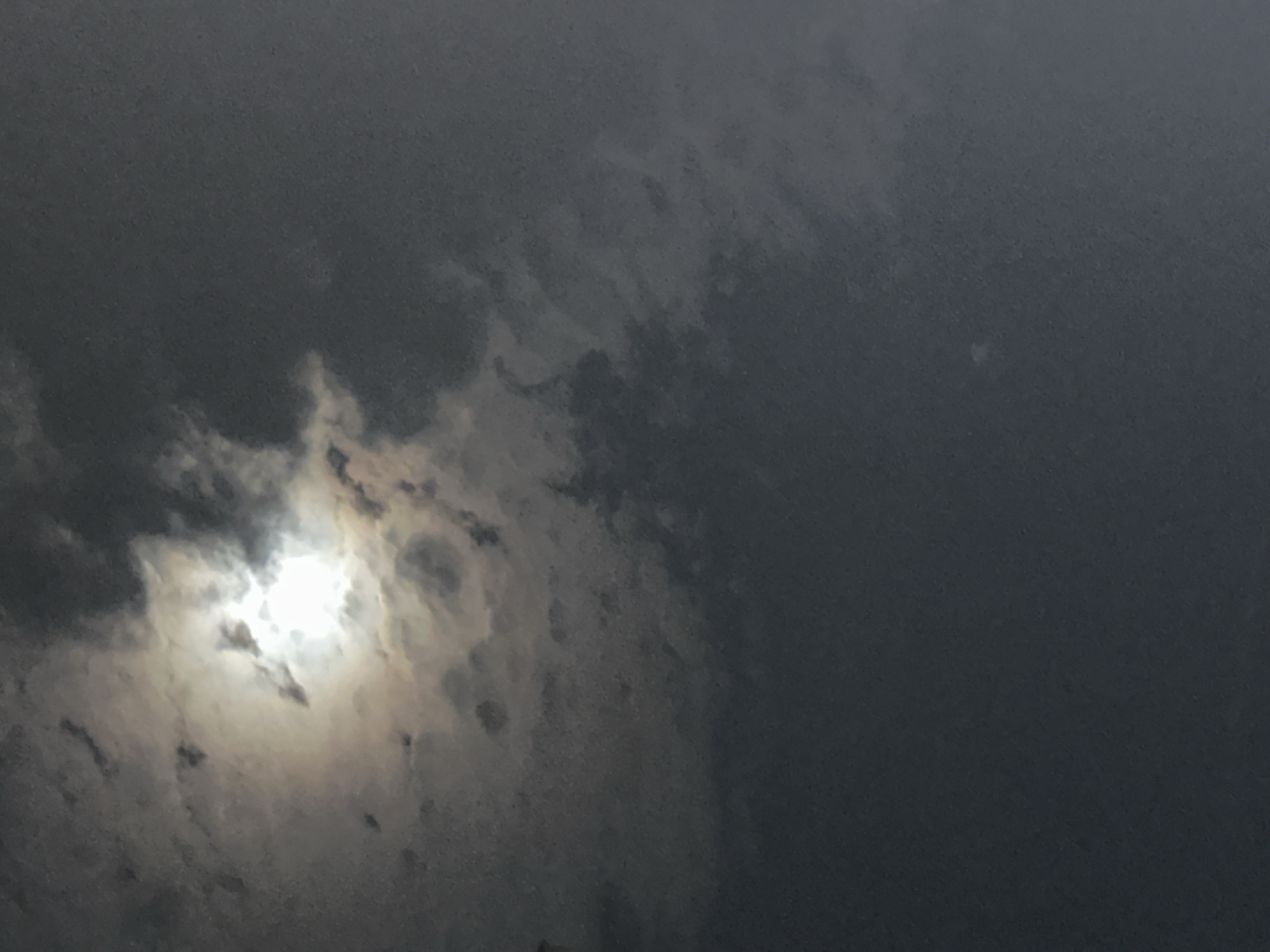 The image is a photograph of a cloudy sky with the sun partially visible behind the clouds. The sun appears bright and diffused by the clouds, creating a luminous glow. The clouds themselves are varied in texture and density, with some areas appearing dark and heavy, while others are lighter and more wispy. The overall mood of the image is somewhat somber due to the overcast conditions. There is no discernible urban environment or specific location within the city of Minsk, Belarus, visible in this image. The photograph appears to be taken from an upward-facing perspective, capturing only the sky. No people, subjects, objects, or actions are identifiable within the frame.