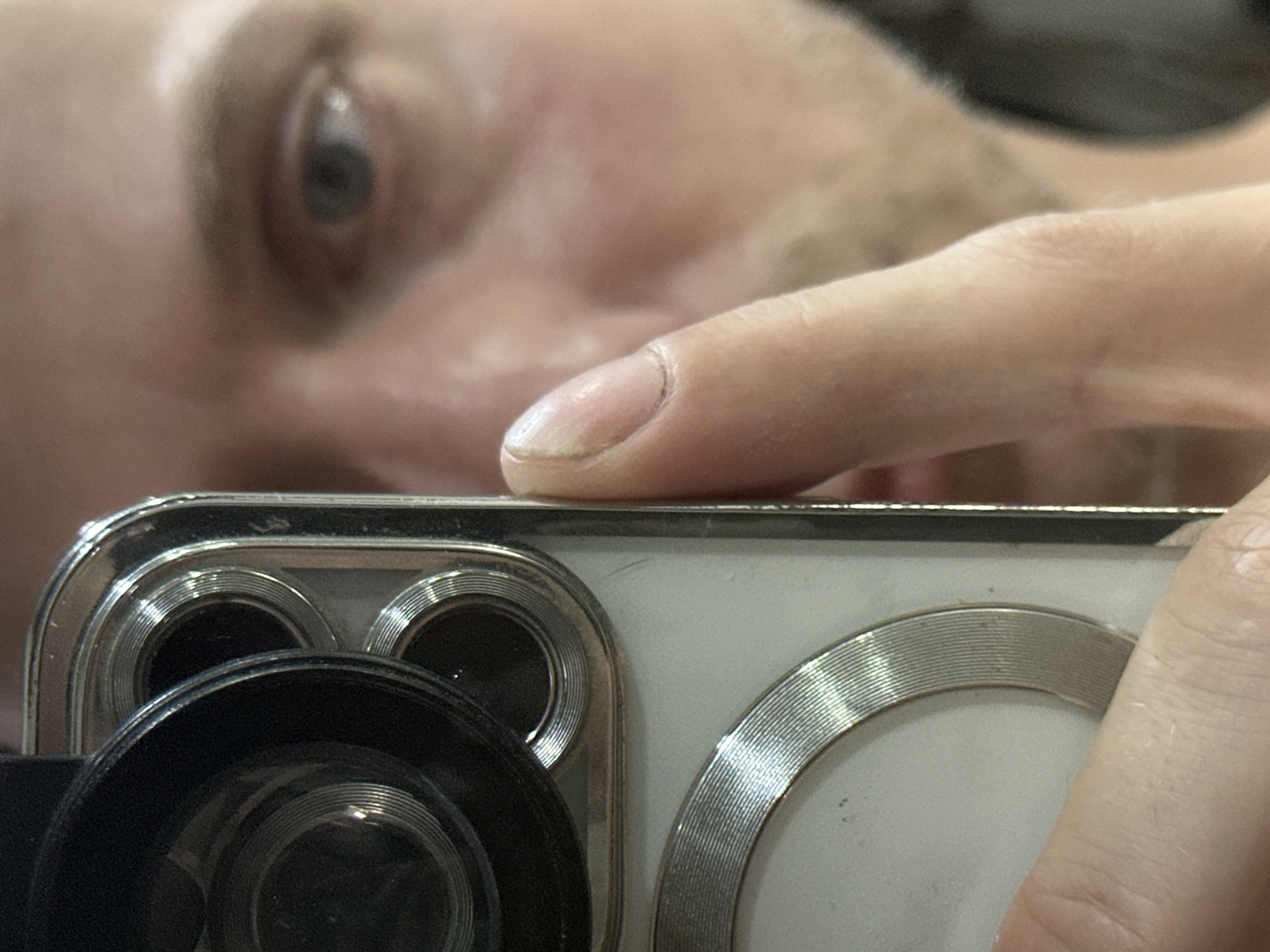 The media file is a close-up, low-angle shot of a person holding a smartphone. The focus is on the smartphone's camera array, which features multiple lenses with textured silver bezels, and the person's hand and finger touching the phone. In the background, a person's eye and part of their face are visible, out of focus. The lighting suggests an indoor setting, possibly during the day, given the natural skin tones. The overall impression is a selfie or a photo taken by someone holding their phone very close. No text is visible in the image. The implied location is Moscow, Russia.