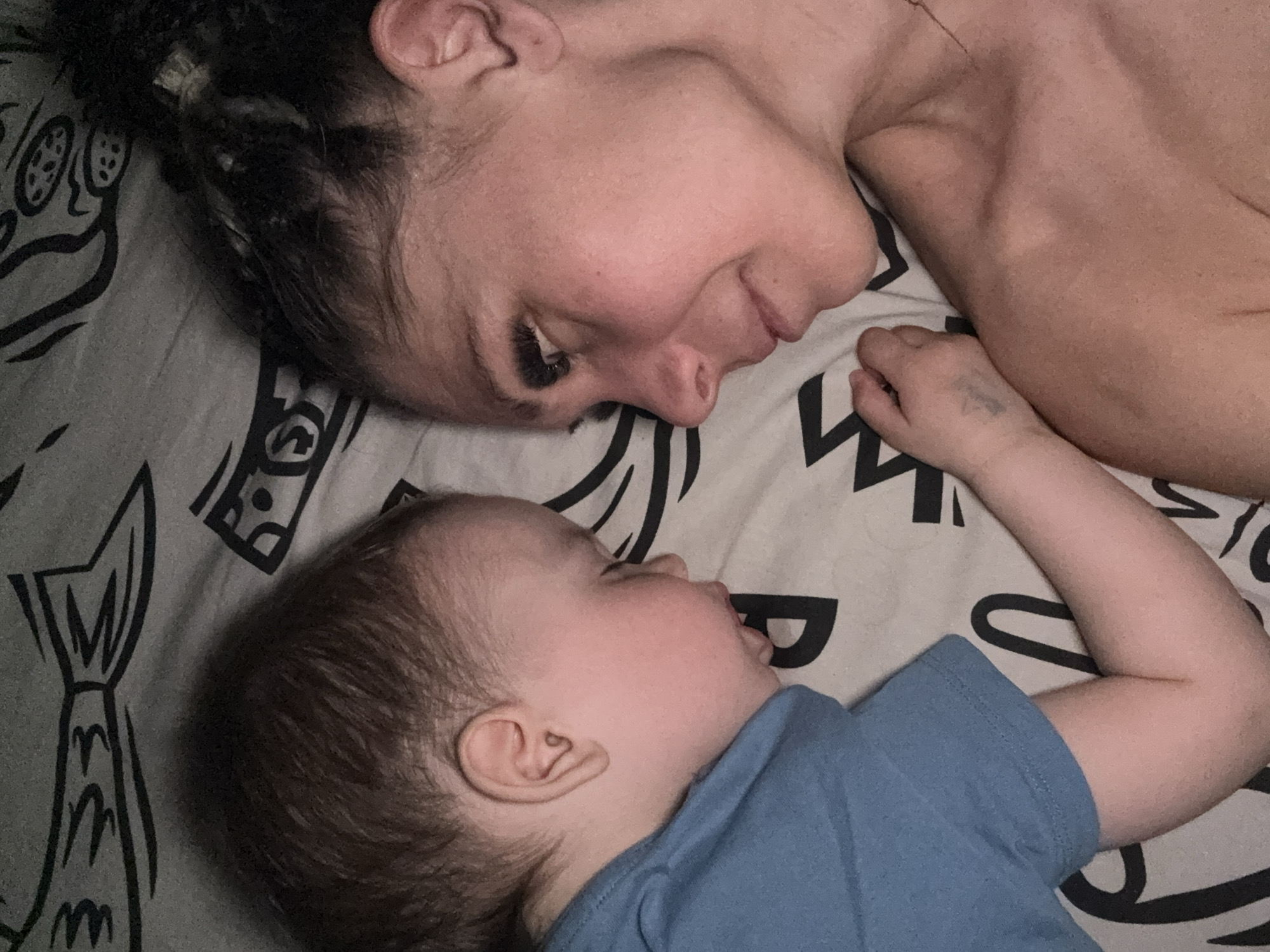 A woman and a baby are lying face-to-face on a patterned bed. The woman is fair-skinned with dark hair pulled back, and she is looking down at the baby with a slight smile. The baby, wearing a blue shirt, is lying on his/her side and also looking up at the woman. The background consists of a light grey bedding with a repeating pattern of what appears to be fish and abstract designs. The location is not discernible from the image.