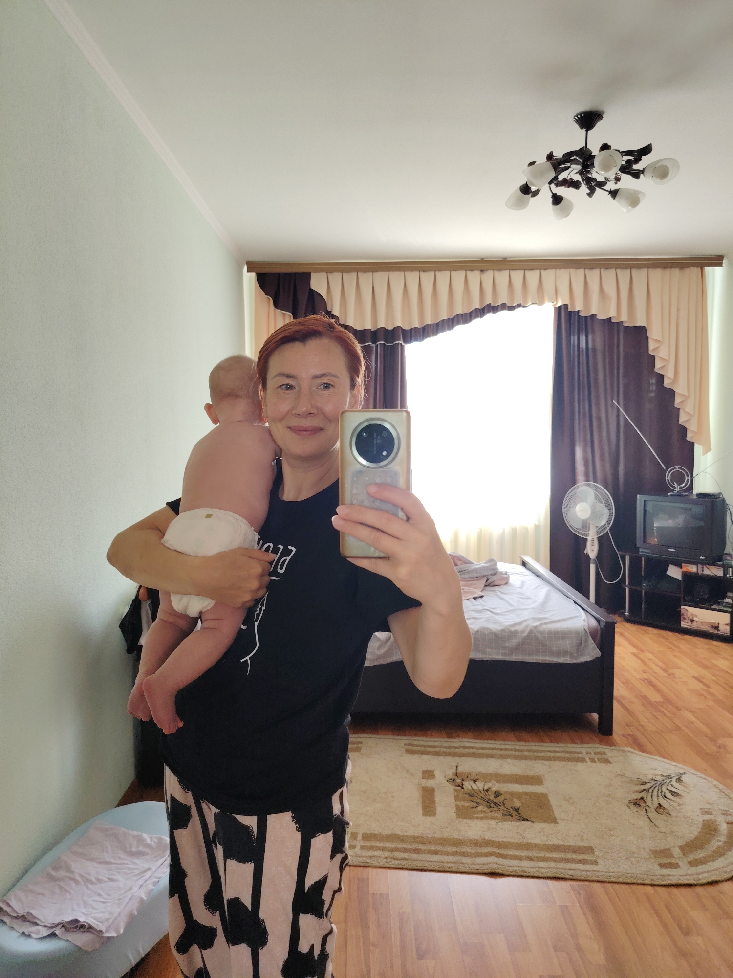 A woman with auburn hair is taking a mirror selfie while holding a baby. She is wearing a black t-shirt and patterned pants. The baby is wearing a diaper and is facing away from the camera. The woman is holding the baby in her left arm. In the background, there is a bedroom with a bed, a fan, a television, and a rug. The room appears to be in an apartment or house. The lighting suggests it is daytime. The location is identified as Konakovo, Russia.