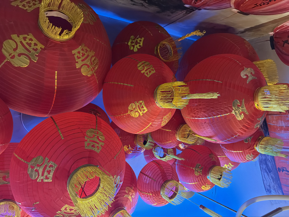 A dense cluster of red Chinese lanterns is depicted hanging overhead in an indoor setting in Canggu, Indonesia. The lanterns, varying in size, are spherical or oblong, constructed from red material, and adorned with gold-colored Chinese characters or auspicious symbols. Each lantern features a yellow or gold fringe and tassel assembly at its base. They are closely arranged, overlapping extensively, creating a decorative canopy. The background primarily consists of a blue-lit surface, possibly a ceiling or wall, with a darker, textured surface visible on the right periphery. No individuals are present in the scene.