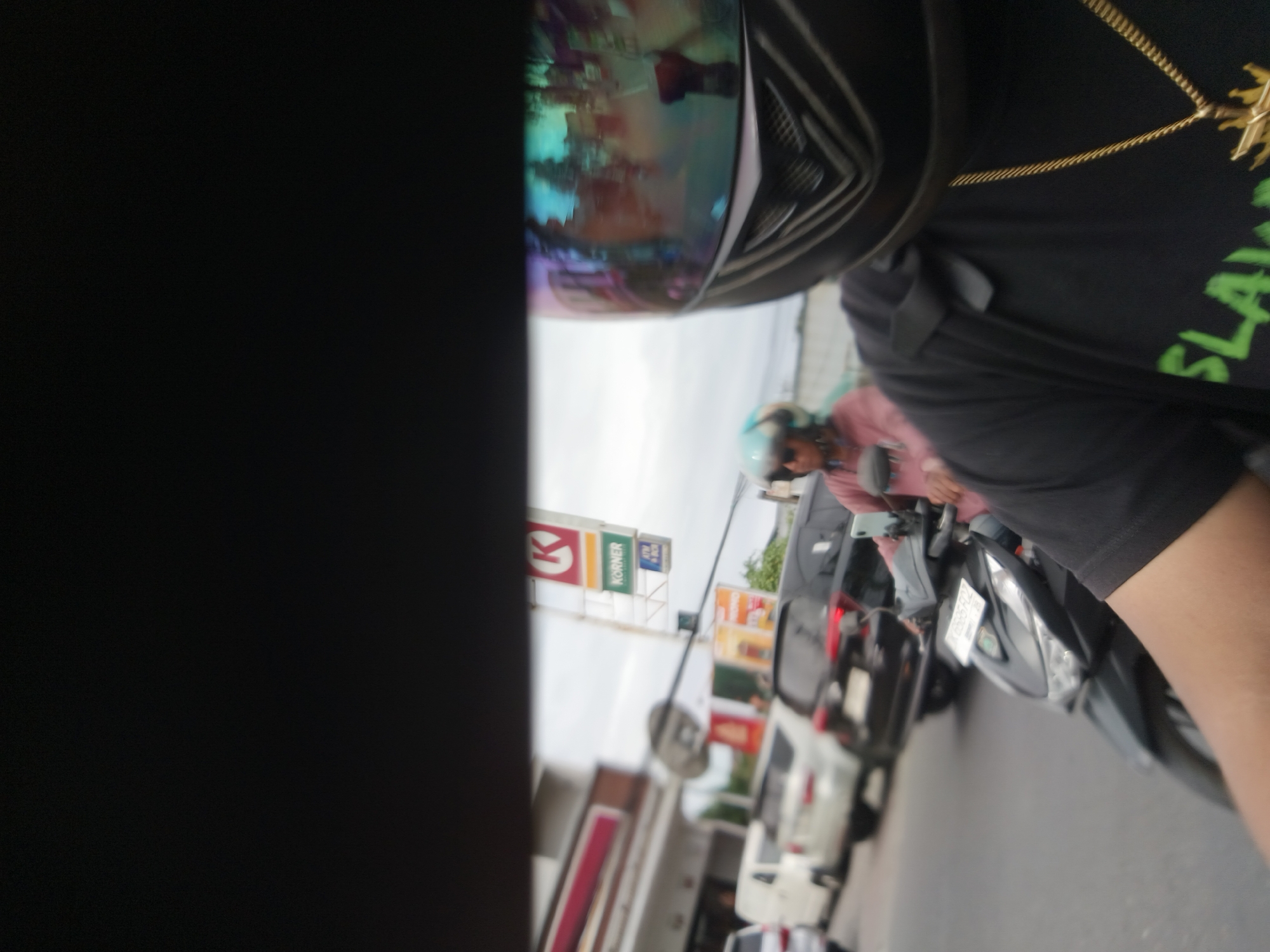 The image depicts a street scene in Denpasar, Indonesia, characterized by traffic and commercial signage. In the foreground, a person wearing a black t-shirt with a gold chain and a helmet with a reflective visor is visible, likely riding a motorcycle. The helmet's visor reflects the surrounding environment, showing a colorful, abstract scene. To the left of the motorcycle rider, a portion of a black object, possibly the rider's clothing or equipment, obscures the view.
Further into the scene, a variety of vehicles, including cars and motorcycles, are present, suggesting moderate traffic. Commercial signs are prominently displayed. One sign, a red "K" logo above the word "KORNER" in green letters, is a recognizable marker of a retail establishment, possibly a convenience store. Other advertisements and signage are visible in the background, indicating a commercial area within the city. The overall environment appears to be an urban street with businesses lining the road. The lighting suggests it could be daytime.