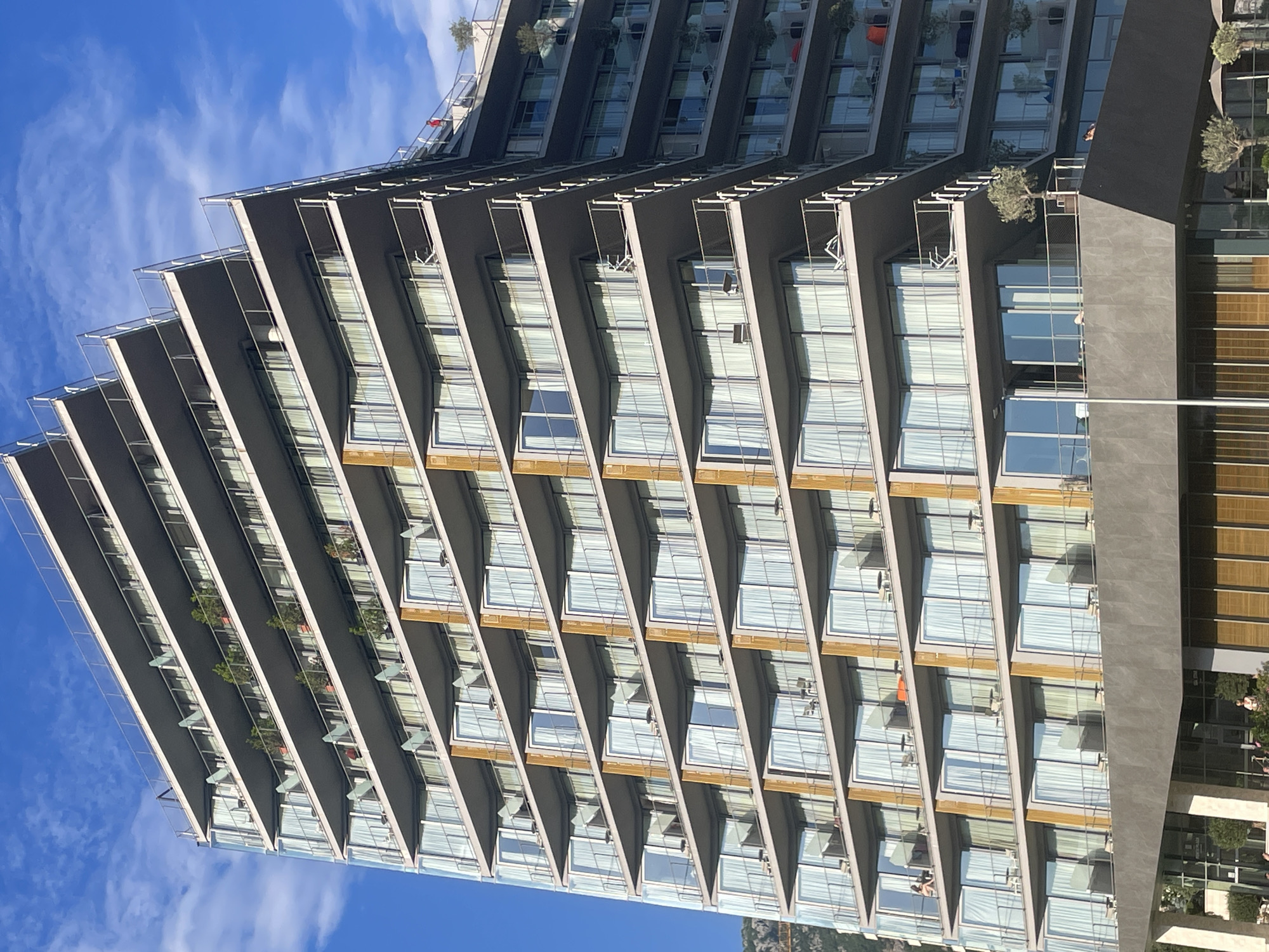 A high-angle view of a modern apartment building in Boreti, Montenegro, with a facade characterized by angular balconies and large windows. The balconies are lined with various plants and, in some instances, white outdoor furniture. The building's color scheme is dominated by shades of gray and brown. Patches of blue sky with light, wispy clouds are visible behind the building.