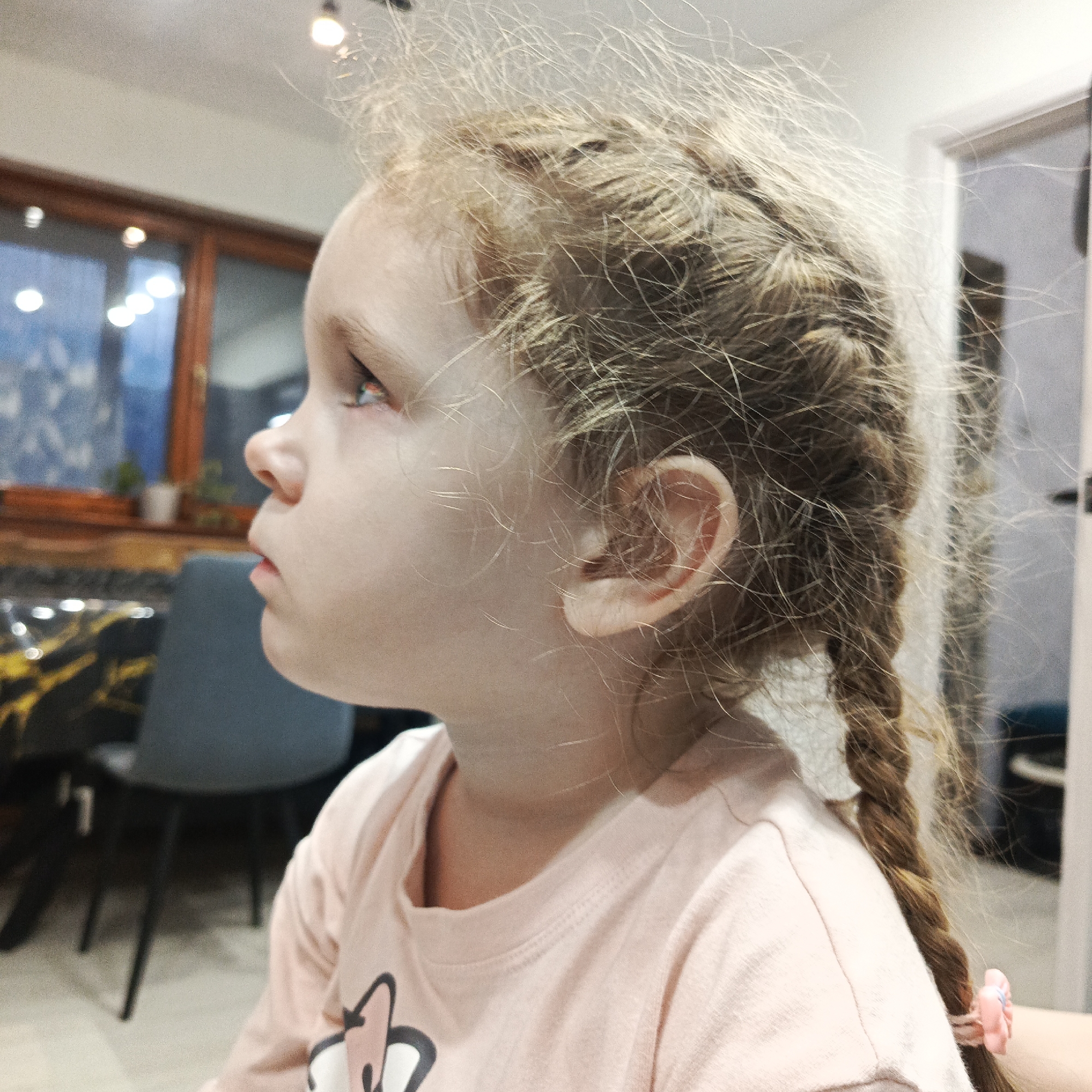 A close-up, profile shot captures a young child, likely a girl, looking upward and to the left with a focused expression. Her fair skin is visible, and her mouth is slightly open. She has light blonde or light brown hair styled in two braids, one of which is visible on the right side of the frame, secured with a small pink hair tie. Numerous fine, wispy hairs are seen standing up around her head, creating a soft halo effect. Her left ear is prominent in the mid-ground.
The child is wearing a light pink, long-sleeved top with a subtle, dark graphic design on the chest area that is not fully legible, possibly a cartoon character or abstract symbol.
The background, largely out of focus, suggests an indoor residential setting in Марковский, Russia. A window with dark wooden frames is visible behind the child, through which blurred outdoor lights and a dark, possibly evening or night, scene can be discerned. The interior features bright, diffuse overhead lighting. To the left, a dark chair with a grey backrest is partially visible, indicating a dining or kitchen area. The overall lighting suggests an indoor setting with artificial light sources, possibly in the evening. No legible text is visible in the image.