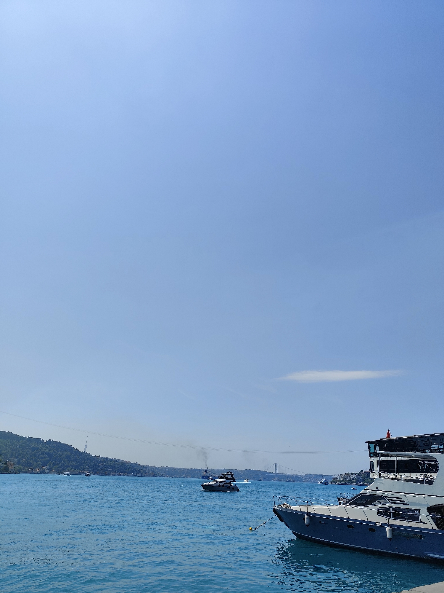 This image captures a wide, bright daytime view of the Bosphorus Strait in Beşiktaş, Turkey, under a vast, clear blue sky. The upper two-thirds of the frame are dominated by the unobstructed sky, which is a vivid cerulean blue with only a single, faint, wispy white cloud visible near the mid-right horizon.
In the foreground on the right, a large, luxurious motor yacht is partially visible, showing its dark blue hull and white upper structure with numerous windows and an enclosed upper deck. A yellow fender or buoy floats beside its bow, indicating it is likely moored.
The deep blue-green waters of the strait fill the lower half of the image, showing gentle ripples and a slight texture from the sunlight. In the mid-ground, several boats navigate the waterway. A dark-colored boat is seen moving towards the left, creating a visible white wake behind it and emitting a plume of dark exhaust smoke into the air. Further back and to the right, another larger vessel, possibly a cargo ship or ferry, also emits smoke as it proceeds along the strait. Other smaller, distant boats are scattered across the water.
The left side of the mid-ground and background reveals a lush, hilly coastline densely covered with green trees and some residential structures nestled among the foliage. In the far distance, a prominent suspension bridge spans across the strait, connecting the two landmasses. The overall impression is one of a sunny, active day on a significant marine thoroughfare. There is no discernible text visible in the image.