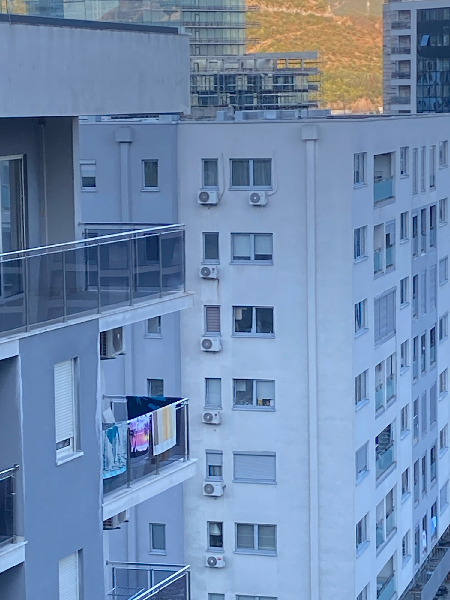 An aerial, medium shot captures the exterior of a multi-story residential building in Budva, Montenegro. The building is predominantly white, with numerous windows of varying sizes and styles. Some windows are covered with blinds, while others offer glimpses into the interior. Several air conditioning units are visible mounted on the building's facade. A balcony with a glass railing extends from one section of the building, with laundry hung to dry on the railing. In the background, other modern buildings are visible, along with a hint of a hilly, treed landscape. The lighting suggests it is daytime, possibly late afternoon, given the warm tones on the distant hills. No people are clearly discernible in the image.