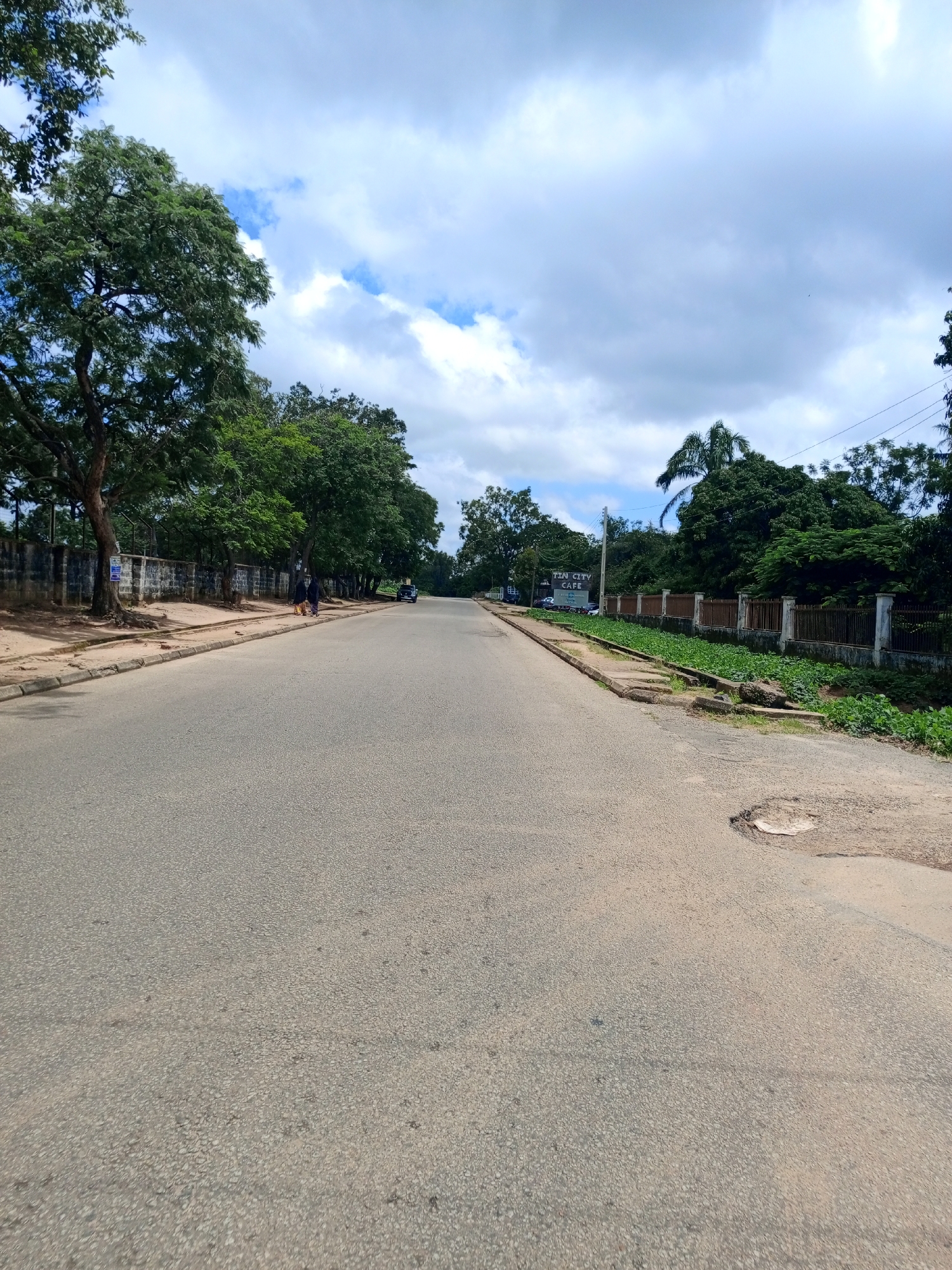 This image captures a view of a road in Jos, Nigeria, under a partly cloudy sky. The road is paved and appears to be a two-lane street, with the perspective looking forward down the road. To the left of the road, there's a sidewalk lined with trees and a brick wall. Further down the road on the left, a sign for "FUN CITY CAFE" is visible, with a dark-colored car parked nearby. Two individuals are walking on the sidewalk, one carrying a yellow bag. On the right side of the road, there's a fenced area with dense green vegetation and a prominent palm tree. The road itself shows signs of wear, including a large pothole on the right side. The overall environment suggests a semi-urban setting with significant tree cover.