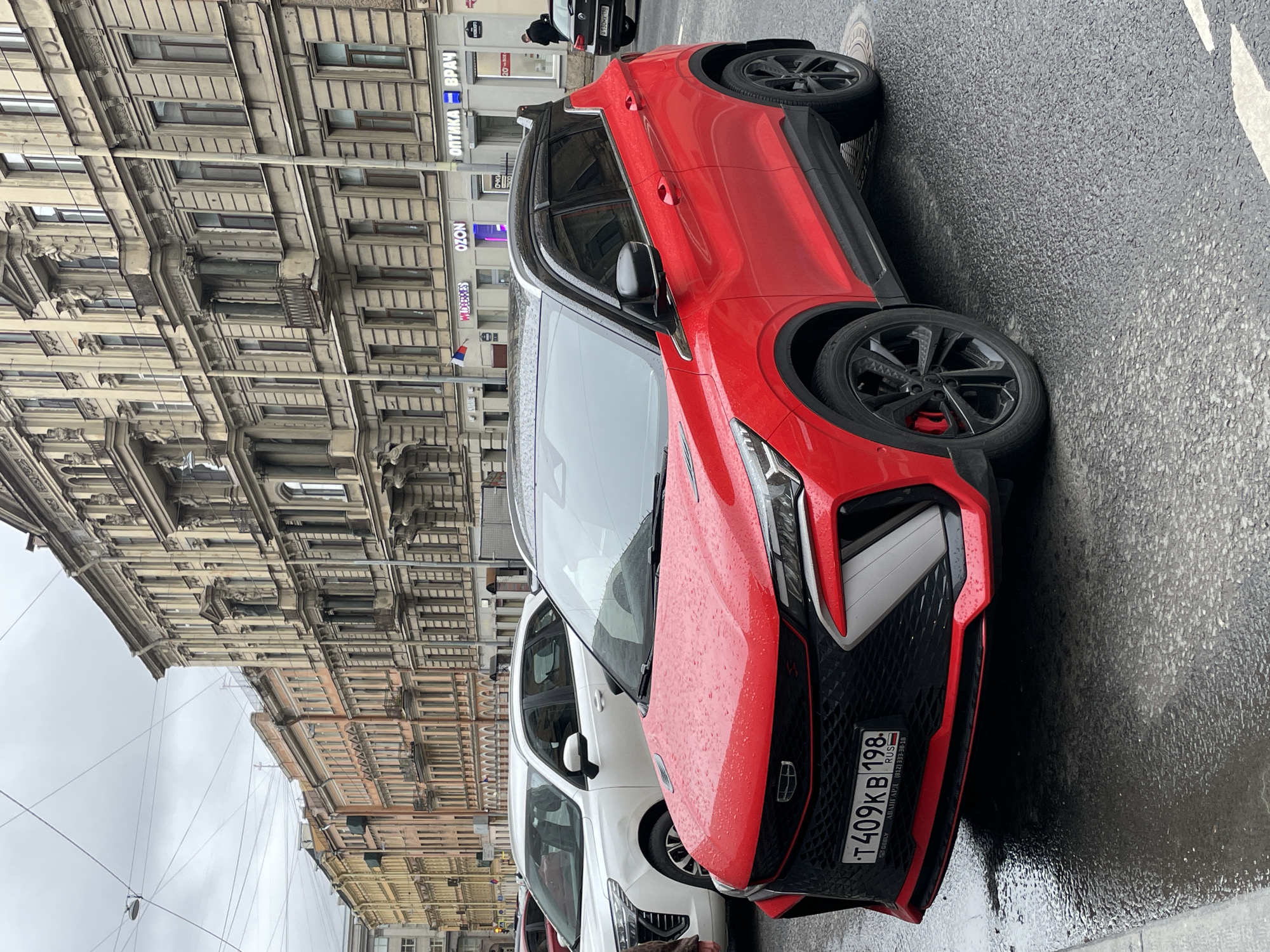 The image captures a red Geely Coolray car parked on a wet street in Saint Petersburg, Russia. The car, with license plate T409KB 198, is the primary subject, with a focus on its front end. The rain-slicked asphalt reflects the surroundings, enhancing the wet environment. Several buildings are visible in the background, typical of Saint Petersburg's architectural style, with multiple windows, detailed facades, and varying heights. Street signs displaying "WIDERRES OZON" and "OПТИКА ВРАЧ" can be identified on nearby buildings, suggesting that the area is a commercial district. Other parked cars are also visible.