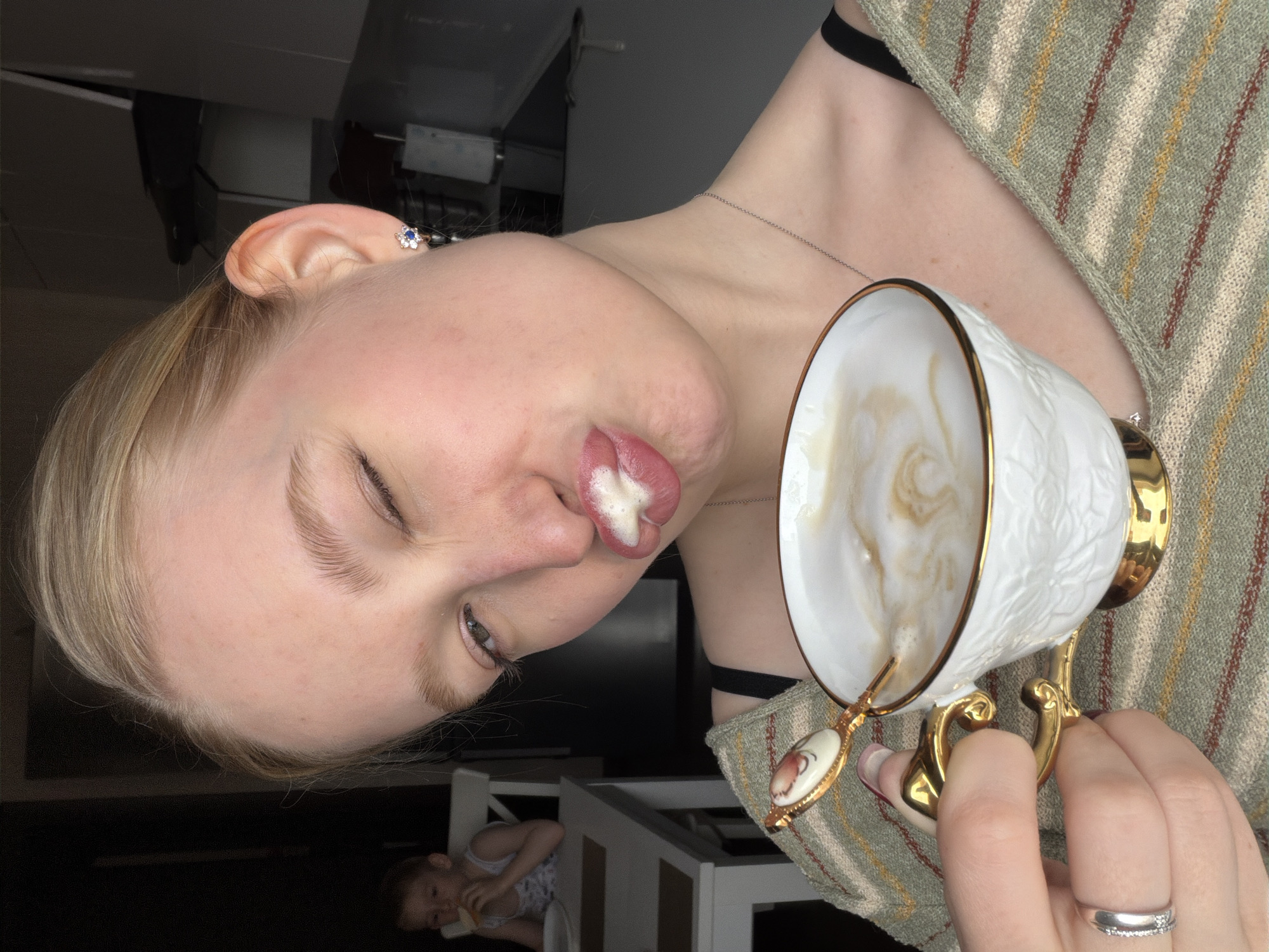 This image, taken in Penza, Russia, is oriented vertically but depicts a scene that would be horizontal if rotated clockwise. It features a close-up of a fair-skinned individual with blonde hair, captured mid-sip from an ornate teacup. The person's head is tilted slightly, and they appear to be winking or squinting their left eye. A notable patch of white foam from the beverage clings to their upper lip.
They are wearing a light green knitted garment with striped patterns in cream and reddish-brown, and a dark strap (possibly a bra or camisole) is visible. A thin silver necklace is worn around their neck, and a blue, flower-shaped earring adorns their right ear. On their left ring finger, a silver band embellished with small stones is present.
The teacup is white with a gold rim and handle, featuring an embossed or textured design on its exterior. It holds a frothy, light brown and white beverage, suggesting coffee or a latte. A small decorative spoon with a patterned handle rests within the cup.
In the blurred background, a young child is visible, seated at a light-colored wooden table or high chair, seemingly engaged with something in their hands. The overall lighting is dim, indicating an indoor setting, possibly an evening or a dimly lit room. The main subject's winking expression and the foam on their lip convey a casual and playful moment. No visible text is present in the image.