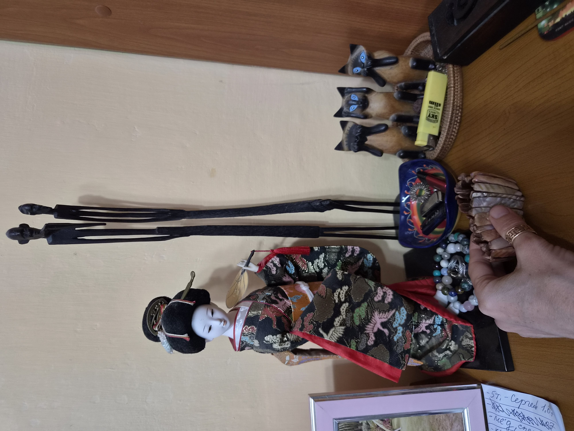 The image depicts a wooden table against a light-colored wall, likely in a residential setting in Arseniev, Russia, as indicated by previous context. A Japanese doll, dressed in a traditional kimono with floral and bird motifs, stands prominently on the table. To the right of the doll are two tall, dark, abstract wooden figures. In front of these figures are three carved wooden cats, each with blue eyes and paws covering their ears, eyes, and mouth, respectively.
In the foreground, a hand is visible holding a bracelet made of shell segments. To the left of the doll, a framed photograph with a pink border is placed. Several strands of beads, including what appear to be various stones and pearls, are scattered on the table near the doll and the hand. A blue, decorative bowl containing items that appear to be a lighter and a pen is situated behind the bracelet and beads. A yellow lighter with "SKY LIGHT" branding is lying on a woven mat in front of the wooden cats. To the far right of the image, a dark, rectangular object, possibly a box or book, is partially visible. A small corner of a paper with handwritten text in Cyrillic script is also present on the left side of the table.